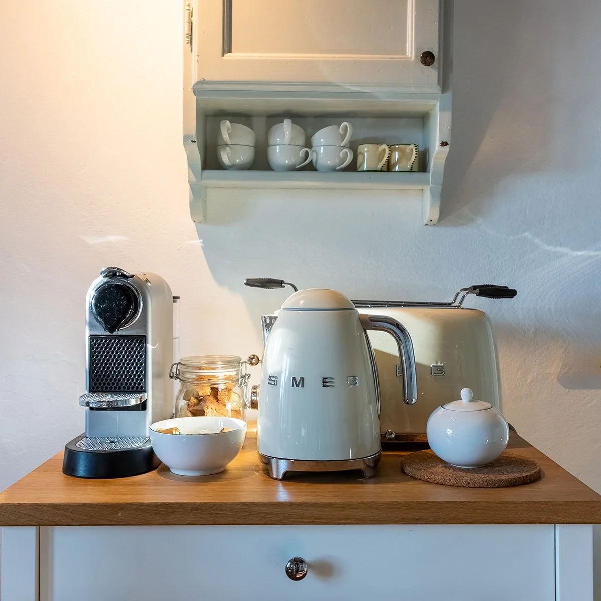 Kitchen counter with a Nespresso coffee machine, a jar of cookies, a bowl, a vintage Smeg kettle, a toaster, and a ceramic container, with a white wall and a wall cabinet in the background.