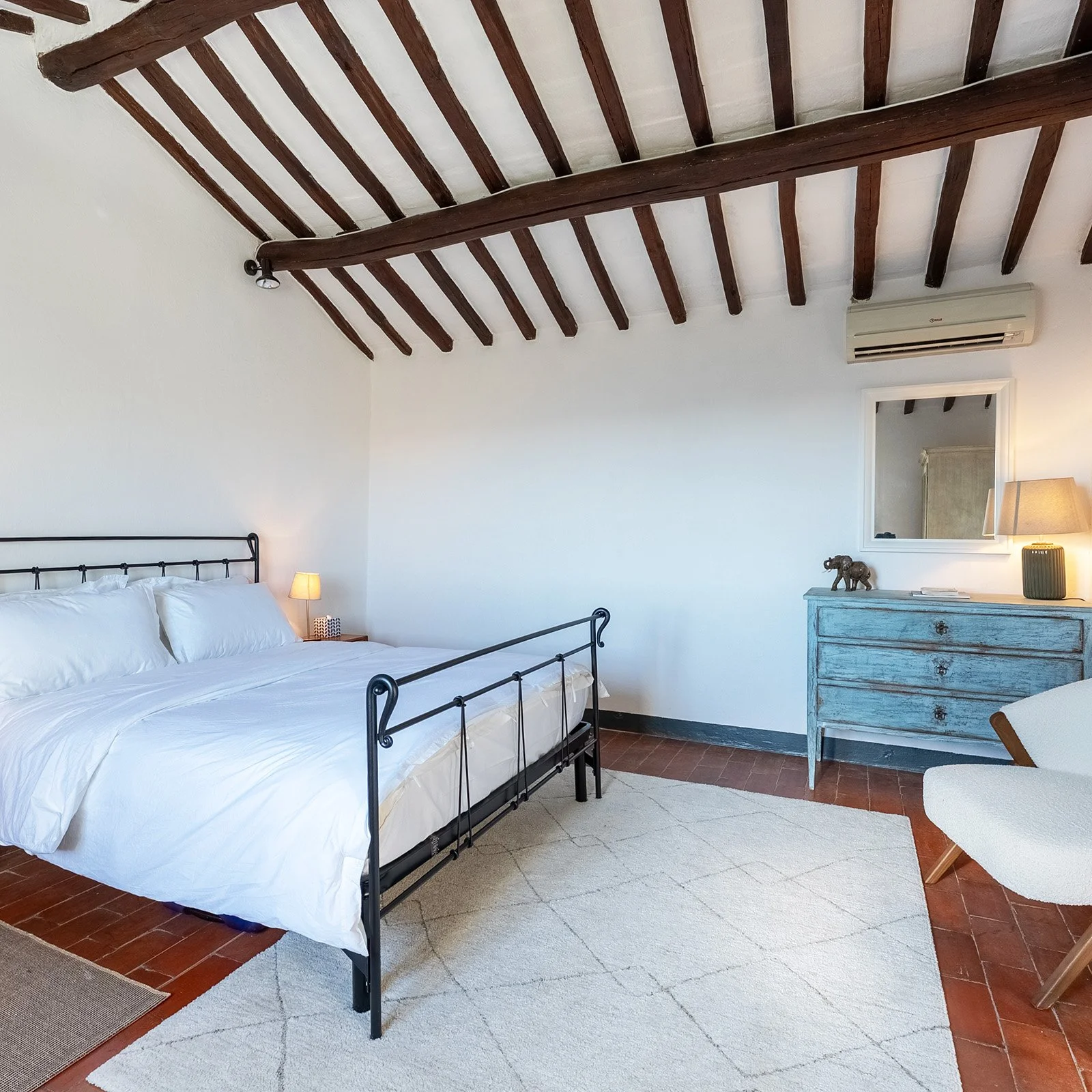 A bedroom with a black metal bed frame, white bedding, a bedside lamp, a blue distressed dresser with a small elephant figurine and a lamp, a mirror on the wall, a white armchair, and exposed wooden ceiling beams.