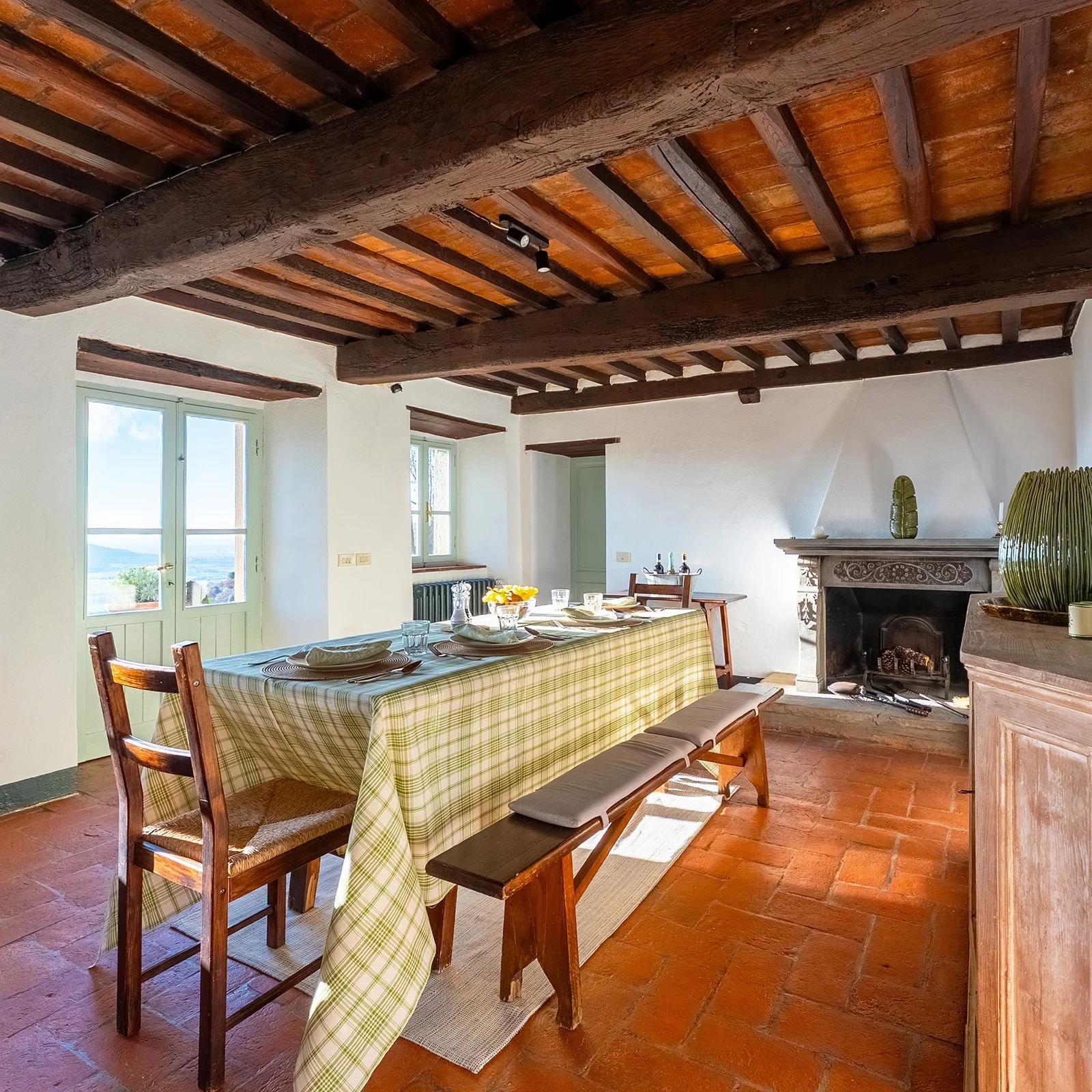 A rustic dining room with a long table covered with a green checkered tablecloth, set with plates, bowls, and glasses. The room has white walls, a wooden beamed ceiling, and terracotta tile flooring. There are large windows letting in natural light, 
