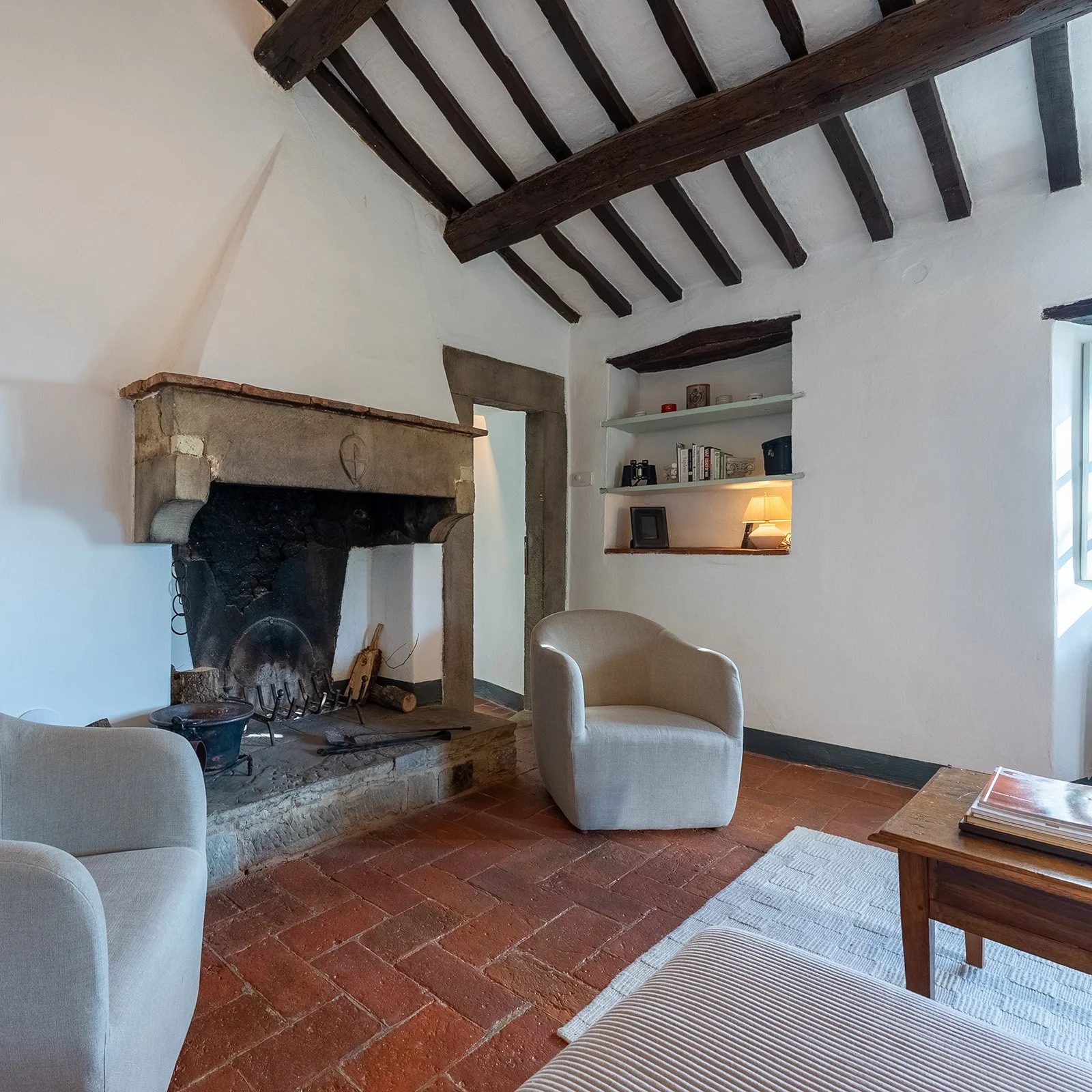 Cozy living room with a large stone fireplace, wooden ceiling beams, white walls, a beige armchair, a wooden side table with books, and built-in shelves with books and a lamp.