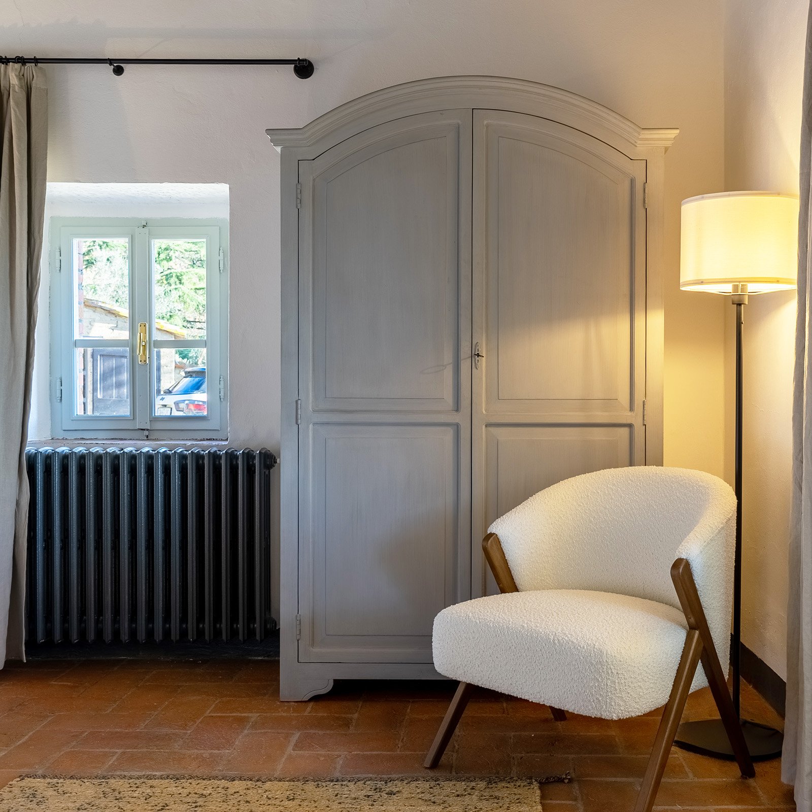 Cozy corner of a room with a white armchair, a tall floor lamp, a white wardrobe, a window with a view of trees and a parked car, and a black radiator.