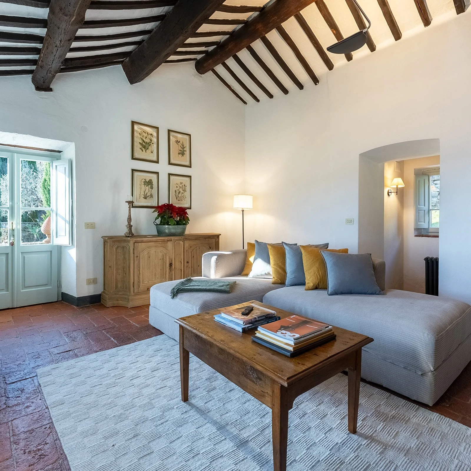 Living room with white walls, exposed wooden ceiling beams, a light gray sofa with yellow and gray pillows, a wooden coffee table with magazines, a wooden sideboard with a poinsettia plant, framed botanical prints, and a floor lamp. There is a window