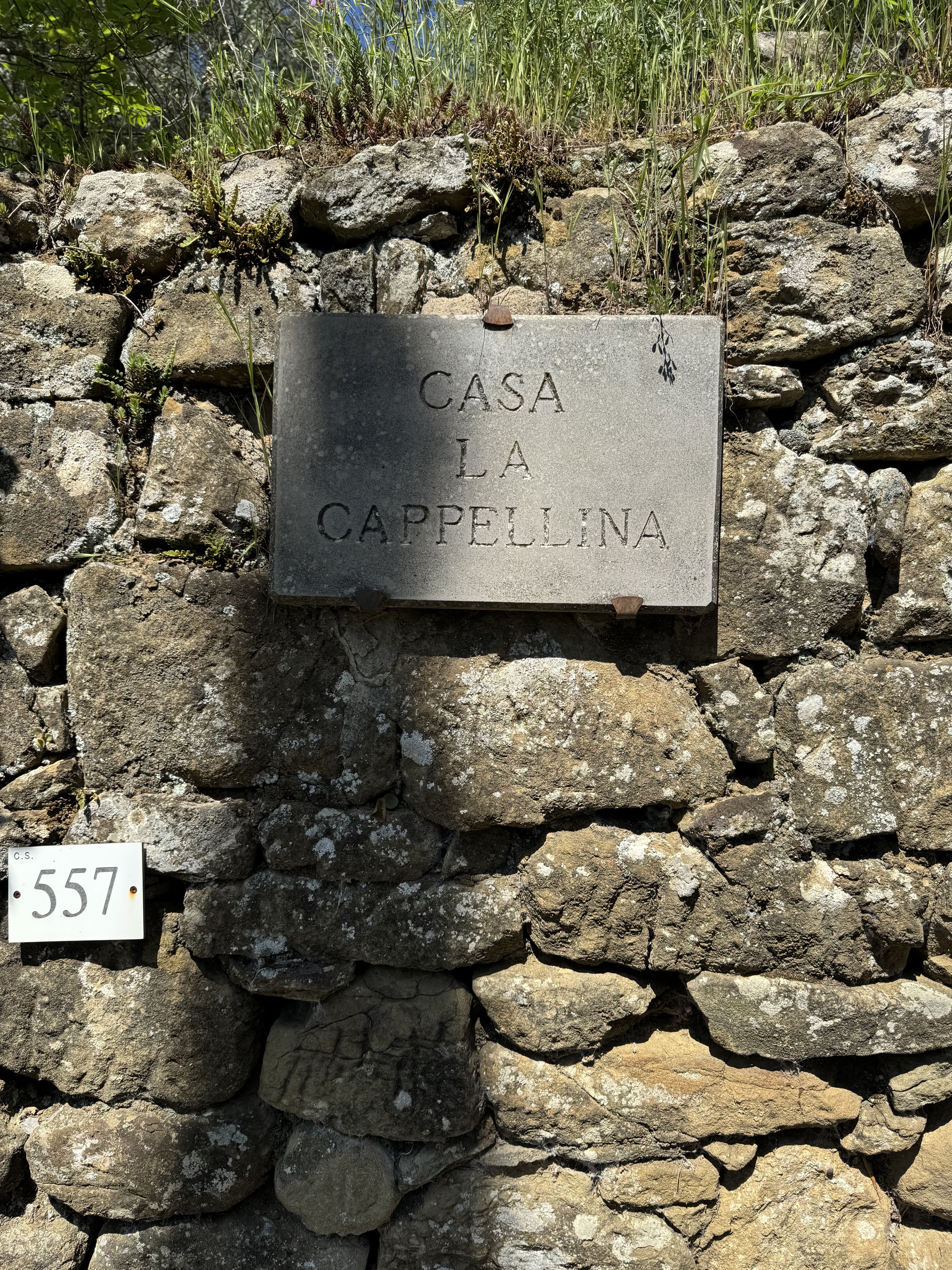 Stone wall with a gray plaque engraved with the words 'Casa La Cappellina' and a small white marker with the number '557' on it.
