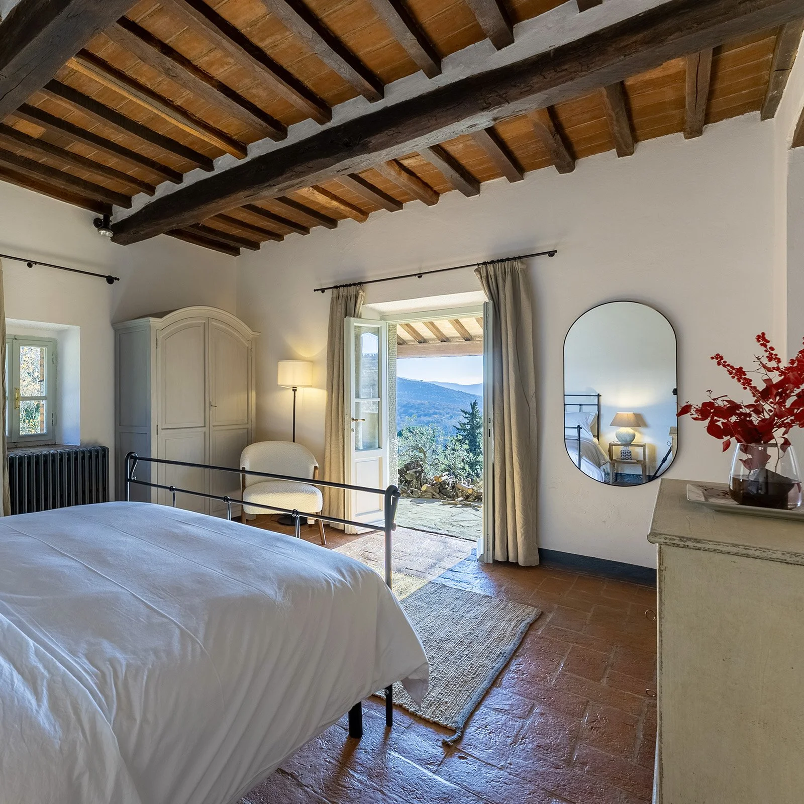 A cozy bedroom with a wrought iron bed, a white wardrobe, a beige armchair, and a standing lamp. There is a large mirror on the wall, a dresser with a vase of red flowers, and an open door leading to a balcony with a view of mountains and trees. The 