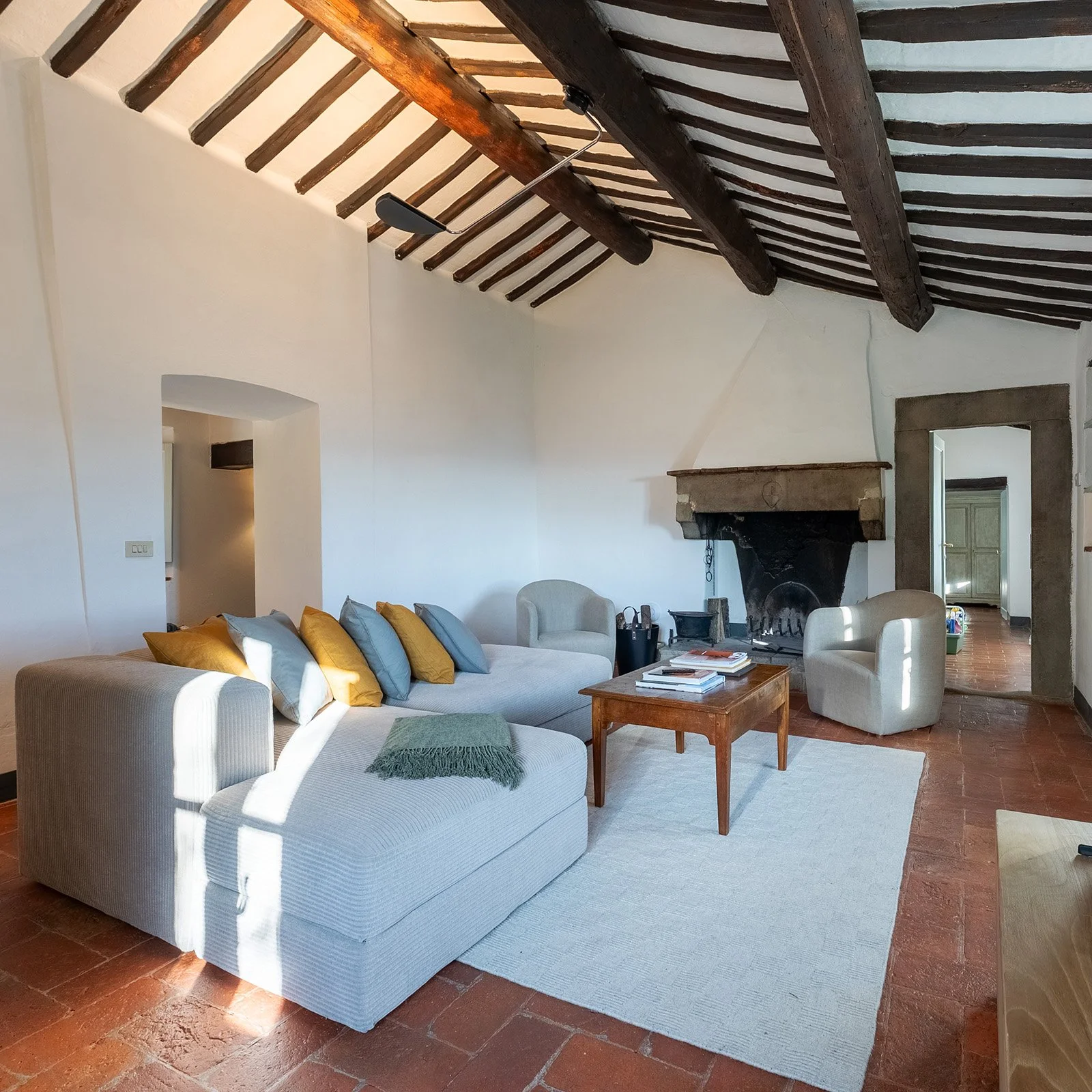 A living room with a high, wooden-beamed ceiling, white walls, a stone fireplace, a large white sofa with yellow and blue pillows, two beige armchairs, a wooden coffee table with books, and a white rug on a terracotta tiled floor.