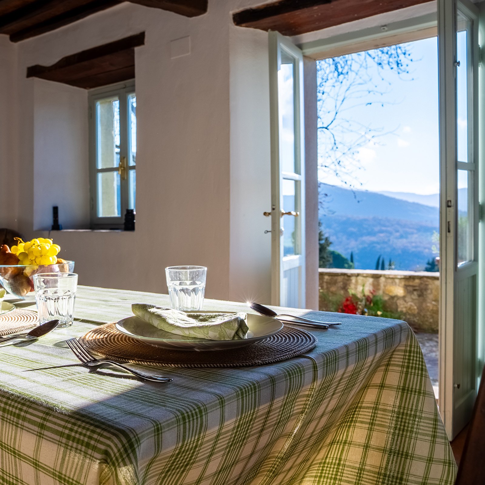 A dining table set for a meal with plates, glasses, silverware, and a napkin, near open doors leading to a scenic outdoor view of mountains and trees.