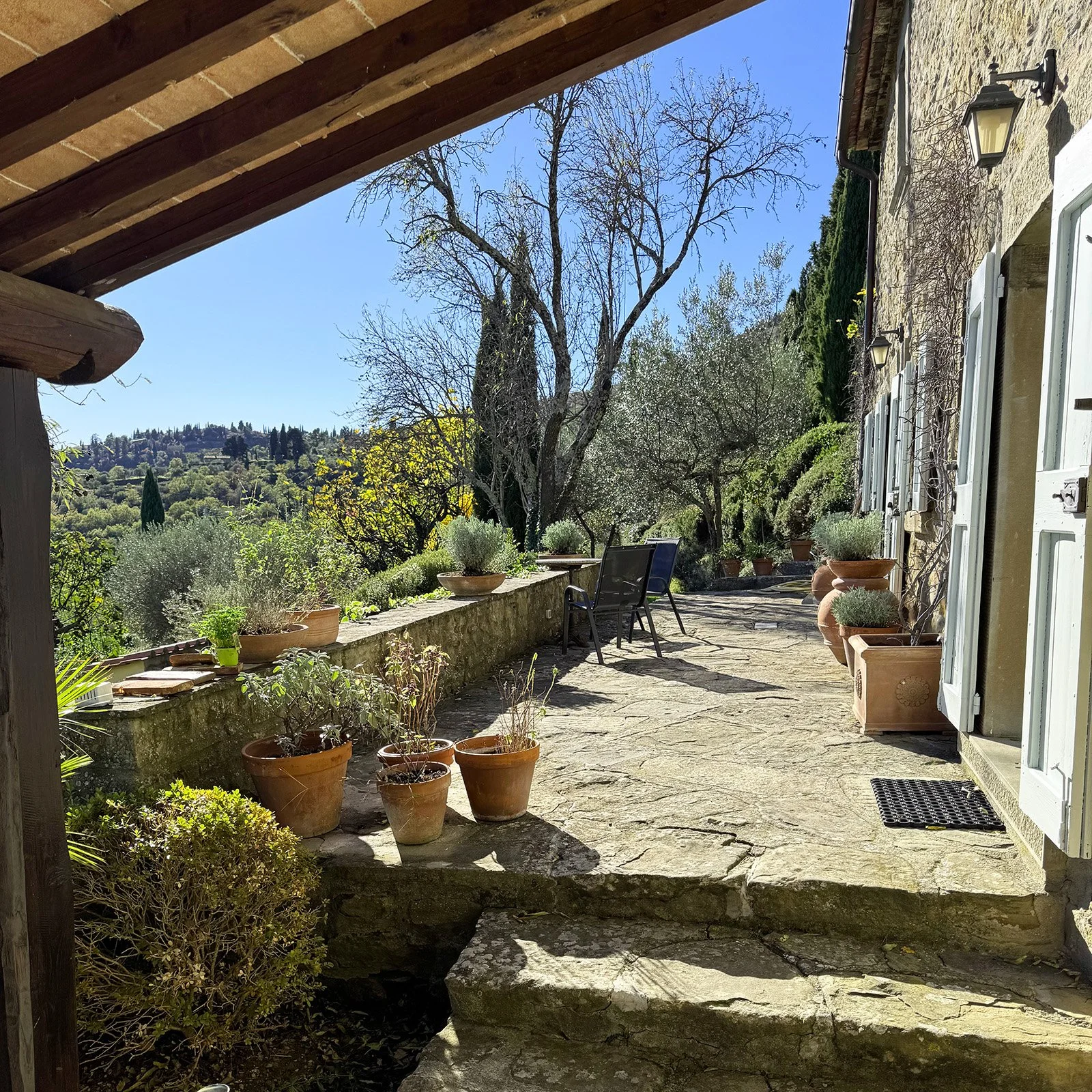Sunlit stone patio with potted plants, a tree with sparse branches, and an hillside view in the background. There are shutters on the windows of the stone house on the right.