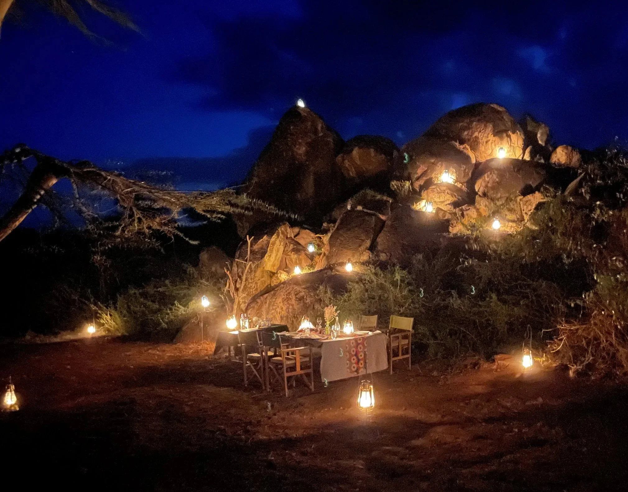 An outdoor dinner setup illuminated by candles and lanterns at night, with large rocks and a tree in the background under a dark sky.