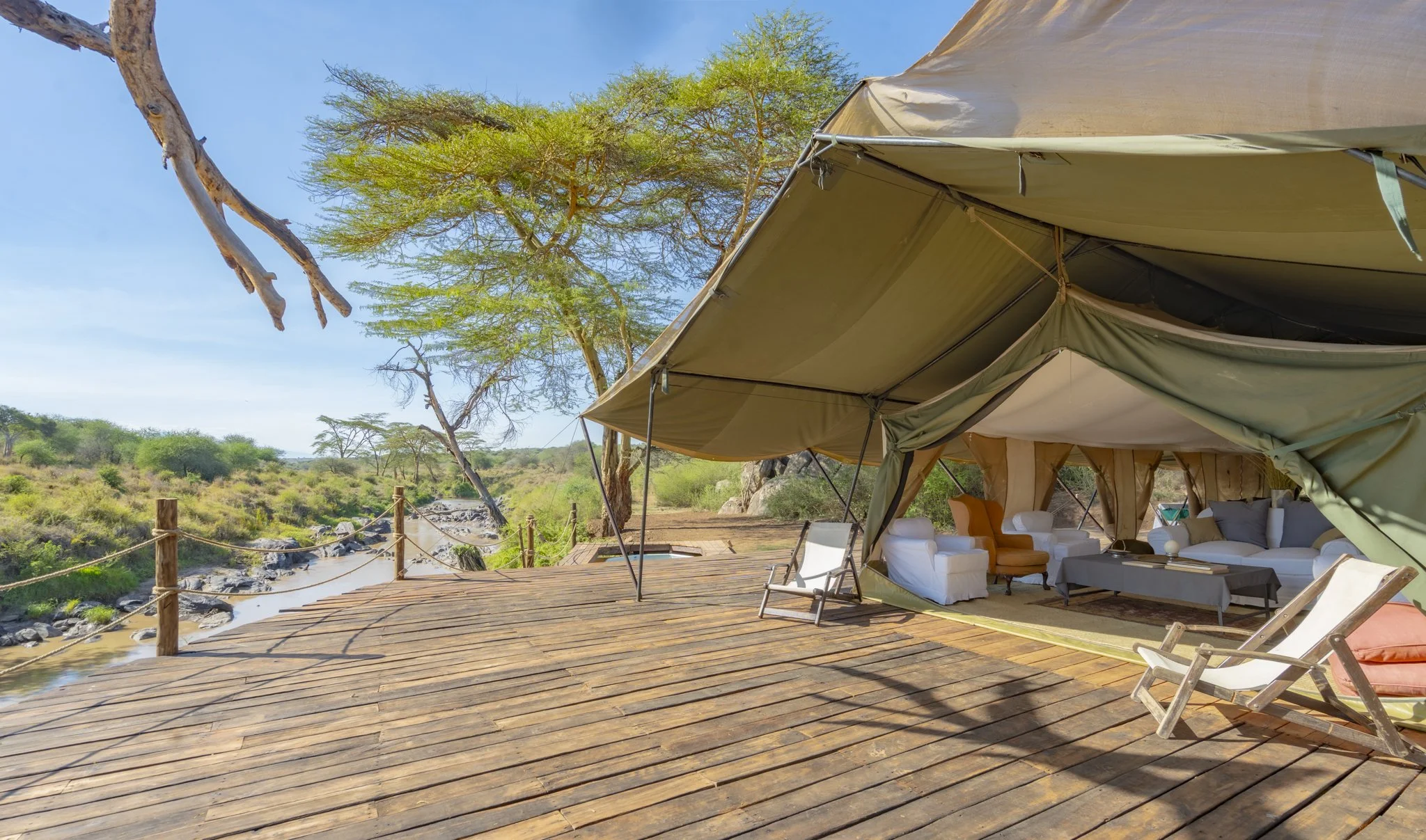 A luxurious outdoor tent with furniture on a wooden deck overlooking a savannah with trees and bushes under a blue sky.