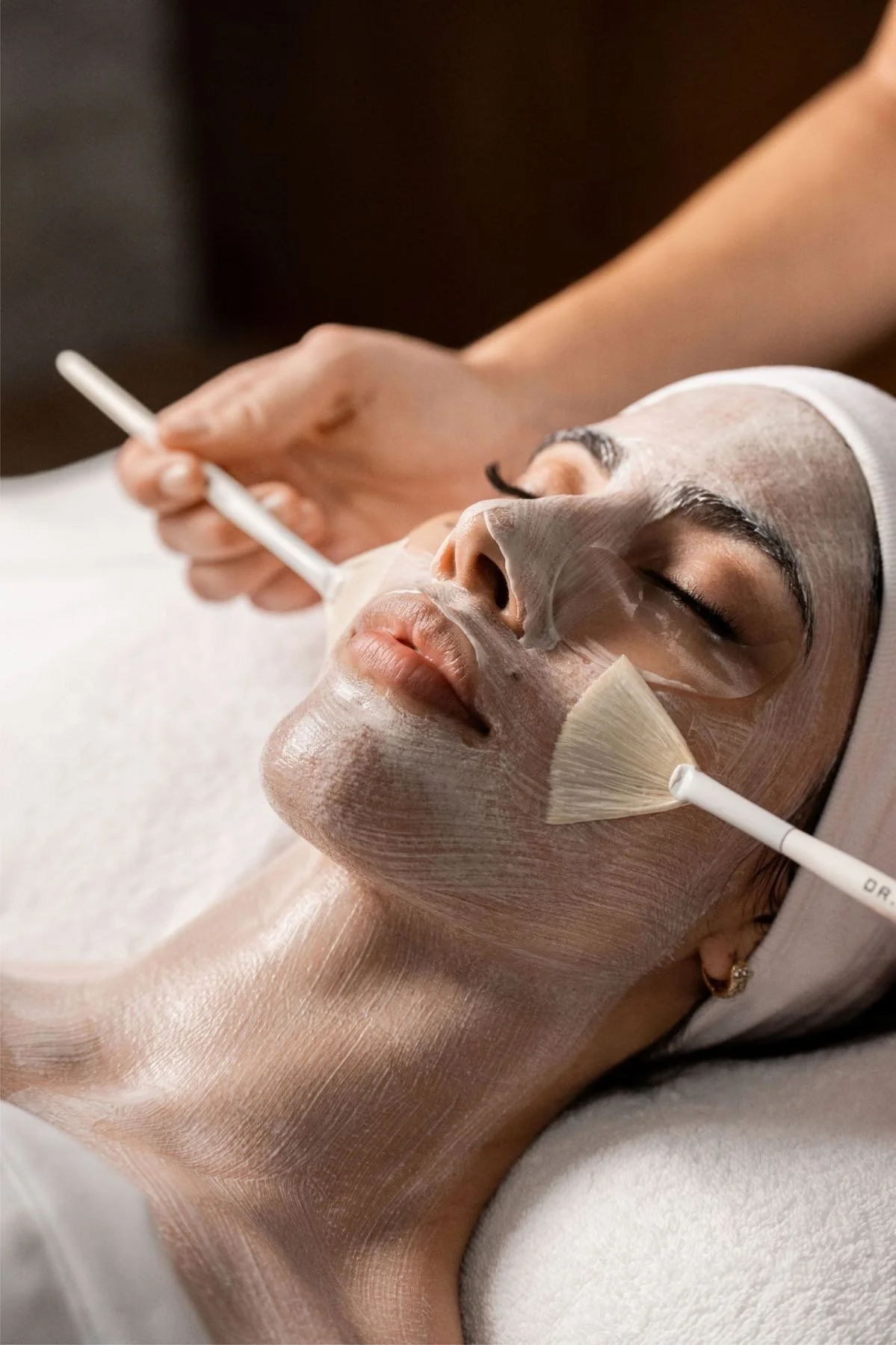 Une femme reçoit un soin facial dans un spa, avec un masque ou un traitement de beauté appliqué sur son visage, alors qu'une esthéticienne utilise un pinceau pour appliquer le produit.