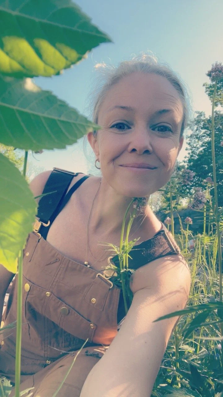 En smilende kvinne med navn Anja Basma tar en selfie blant høye grønne planter utendørs på en solrik dag.