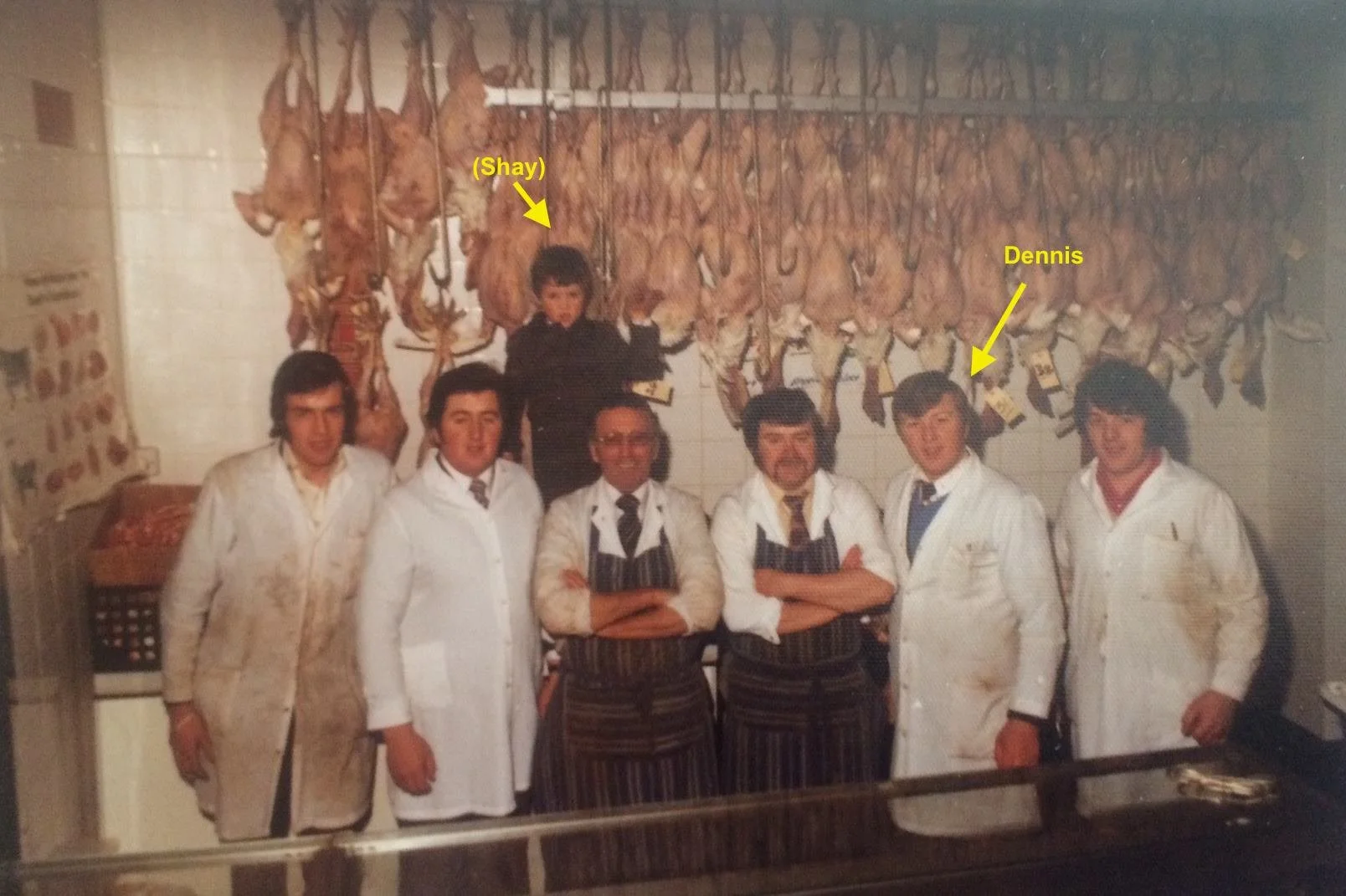 A group of seven men and one young boy in a butchery with hanging animal carcasses on the wall behind them. The men are dressed in white coats, with some wearing striped aprons, and the boy is in dark clothing. The area appears to be a meat shop or b