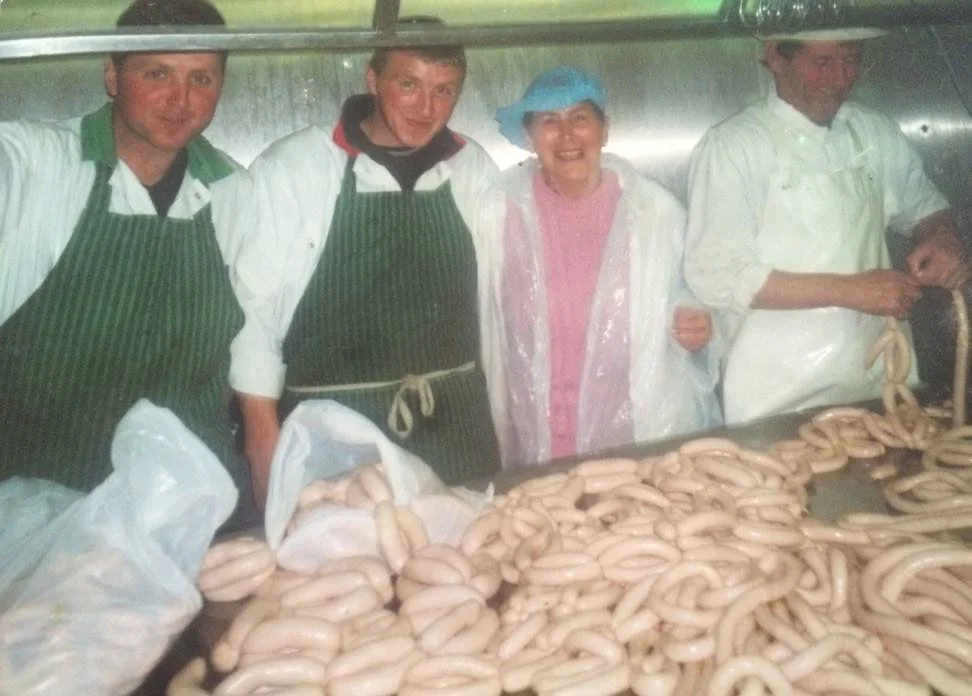 Four people standing behind a table with a large pile of sausages, in a food processing or butchery setting.