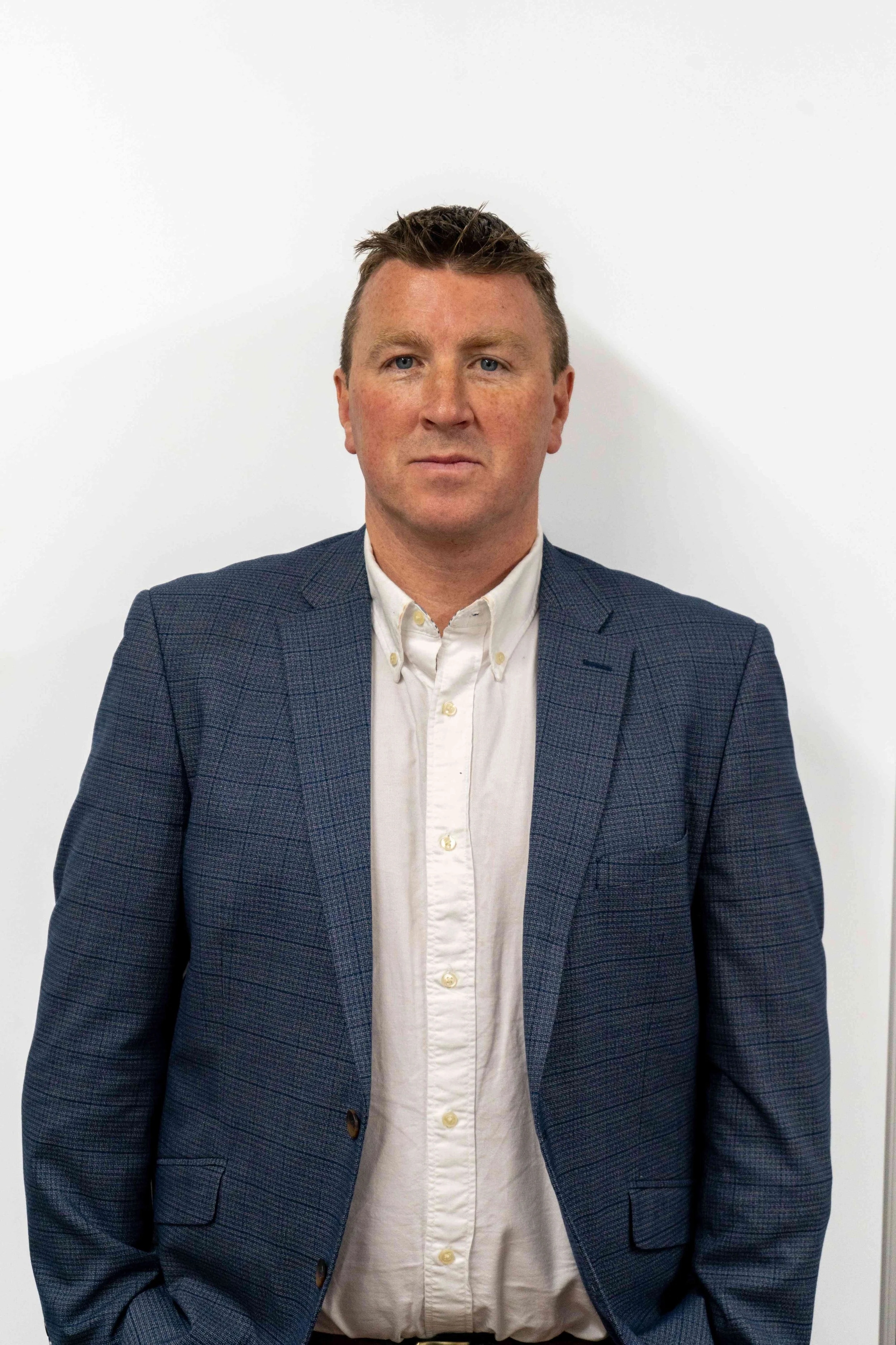 A man with short brown hair, wearing a white collared shirt and a dark blue checked blazer, standing against a plain white background.