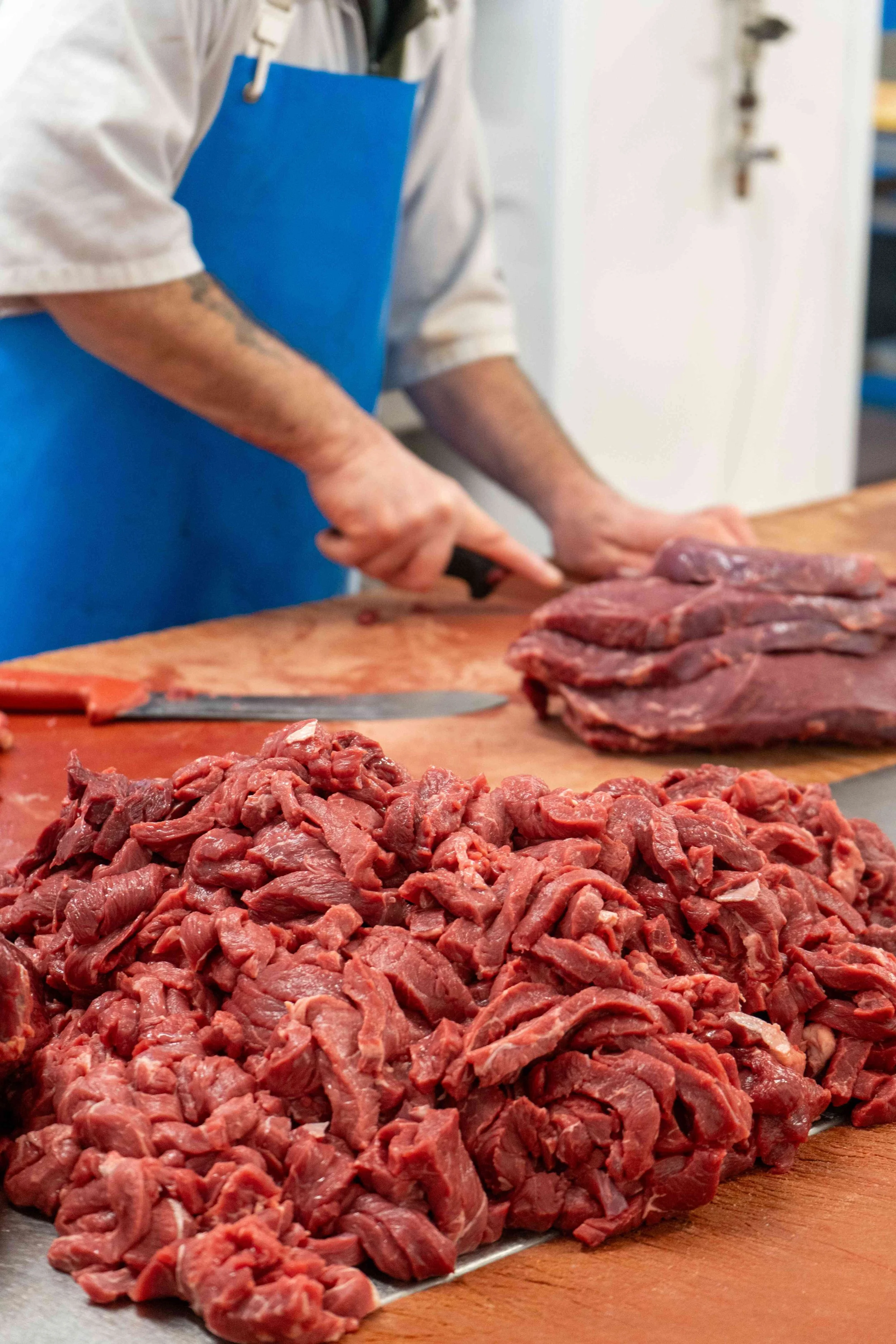 Person in a white shirt and blue apron butchering beef on a wooden table.