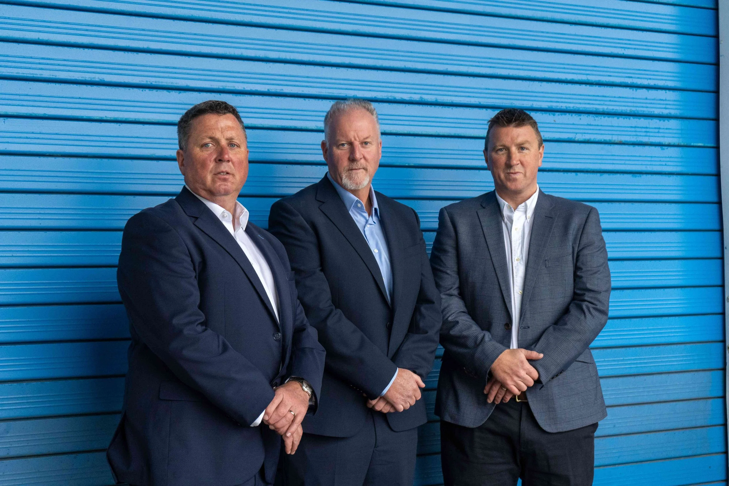 Three men in suits standing in front of a blue metal roller shutter door.