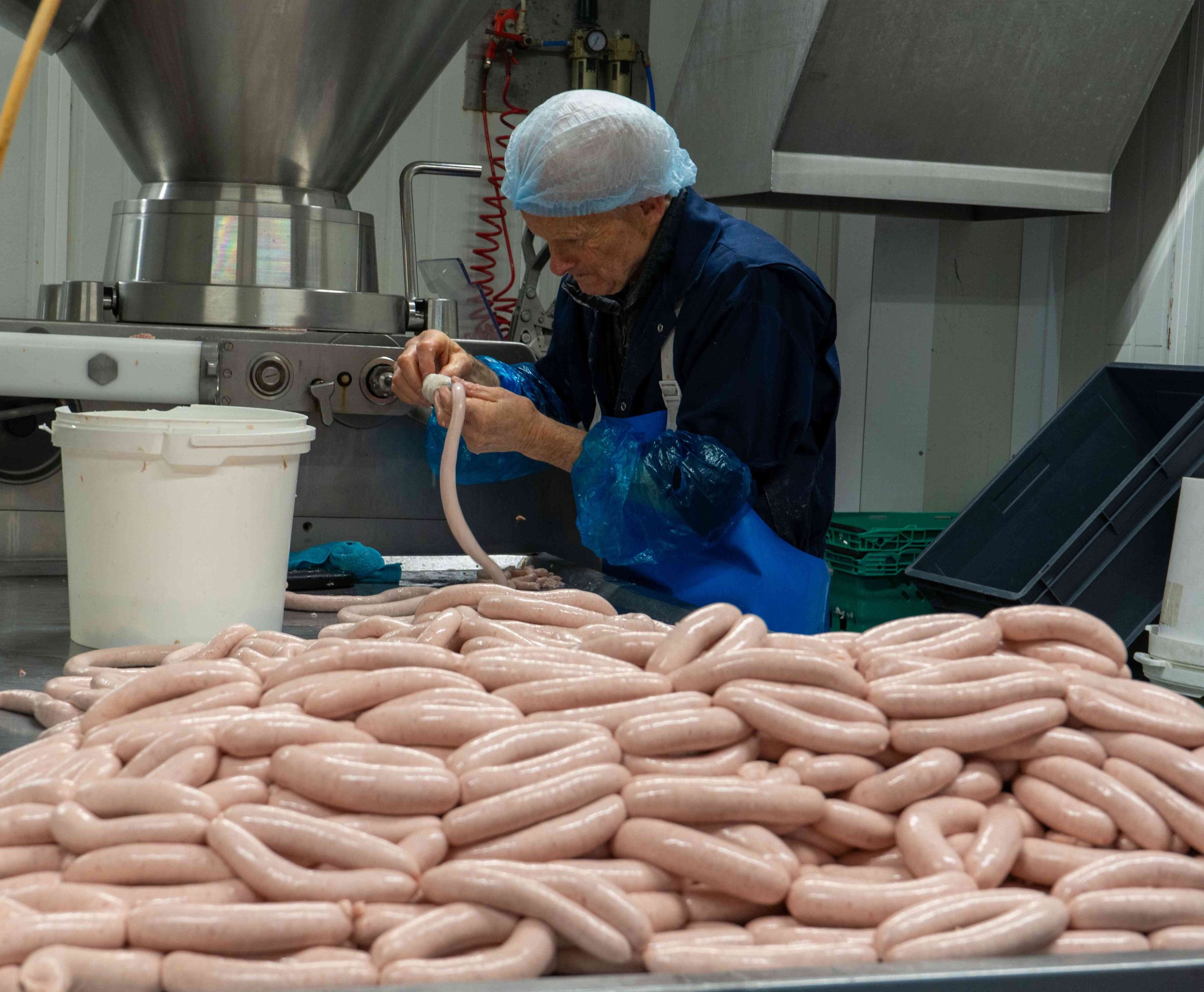 An elderly worker in a hairnet, apron, and blue gloves processes sausages in a meat processing facility, with a large pile of sausages in the foreground.