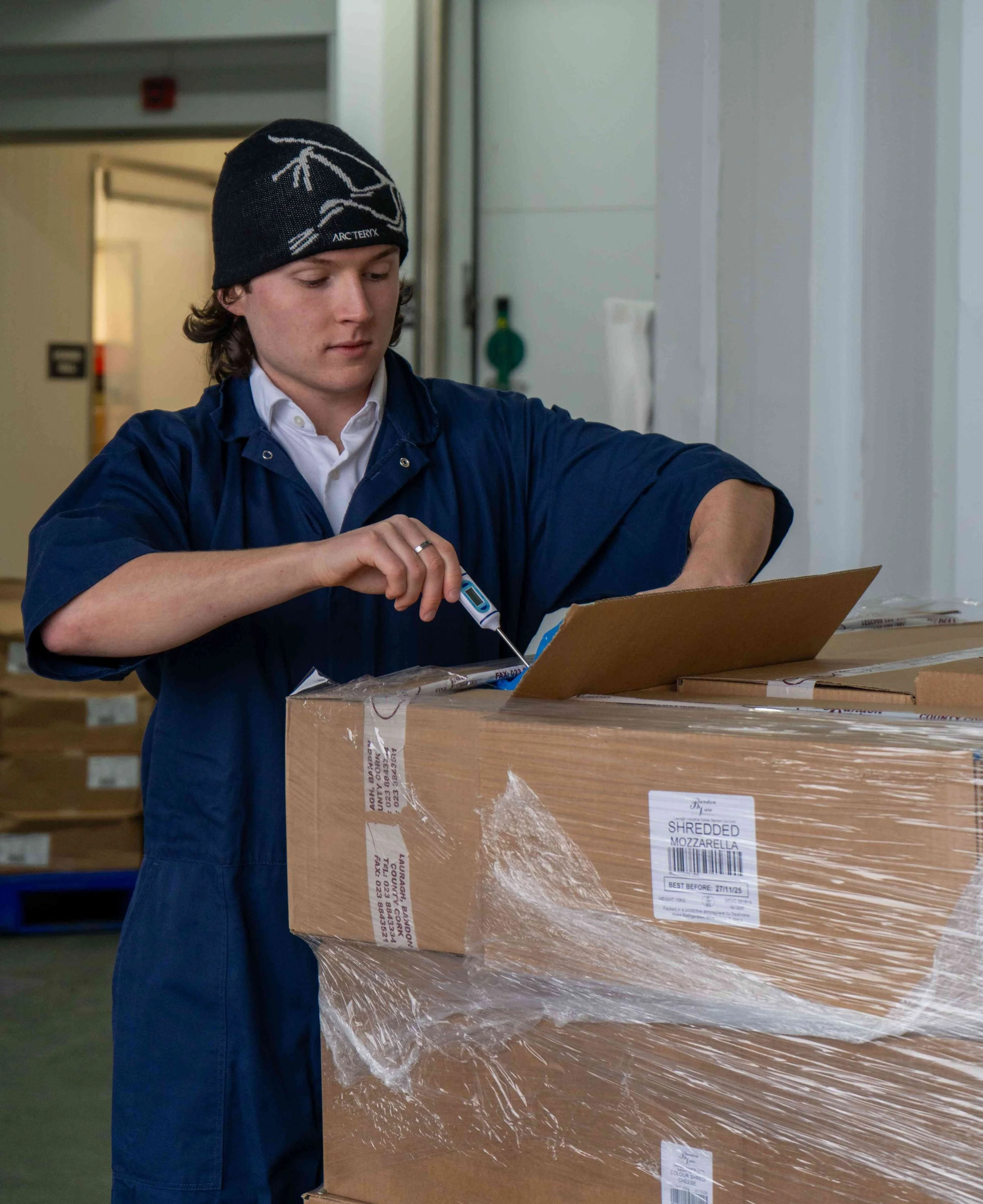 A young man in a blue work coat and a black beanie hat with a white design is inspecting a large cardboard box labeled 'Shredded Mozzarella' with a thermometer in his hand in a warehouse.
