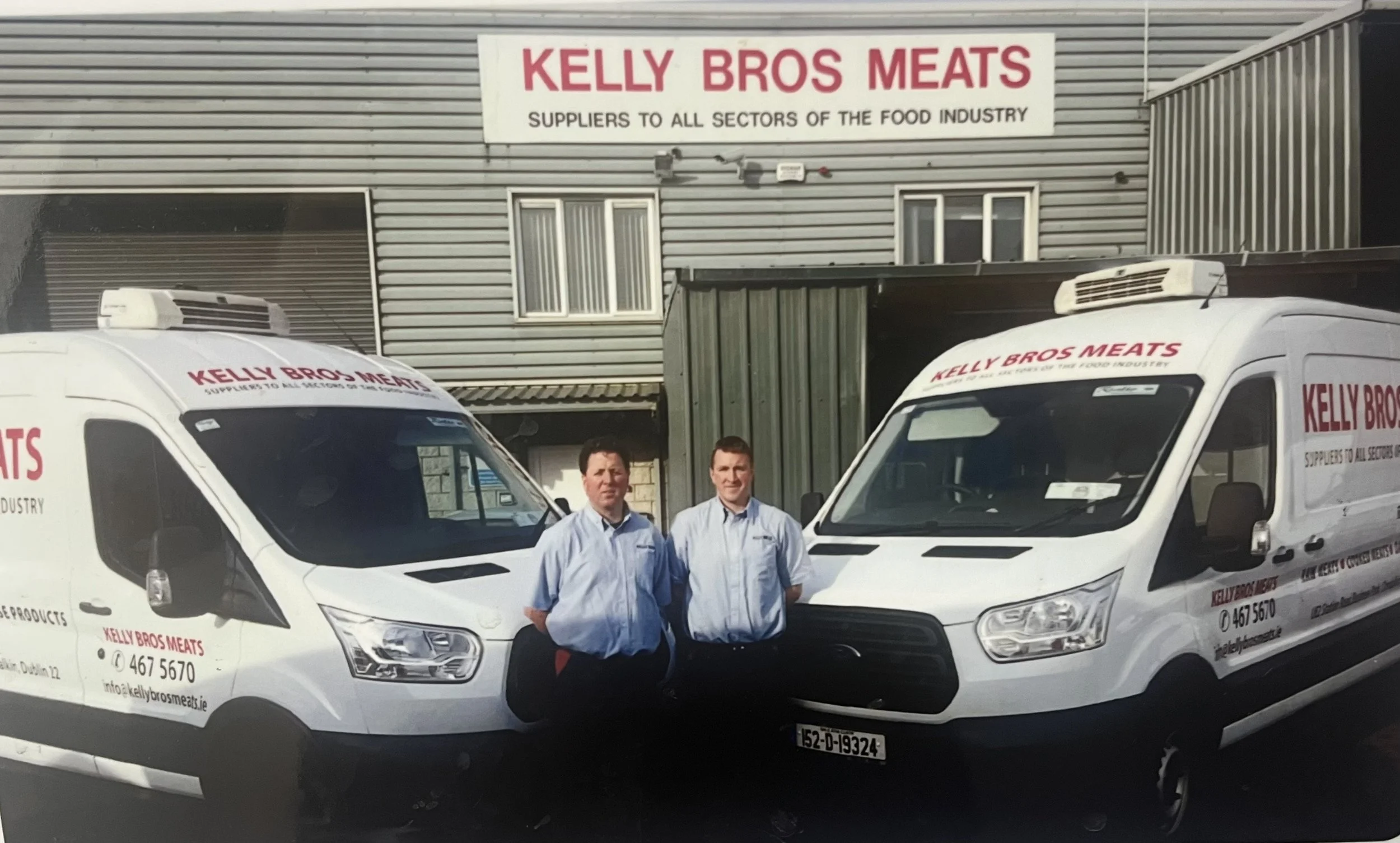 Two men in blue shirts standing in front of white delivery vans with Kelly Bros Meats branding, parked outside a building with a sign for Kelly Bros Meats, a food industry supplier.