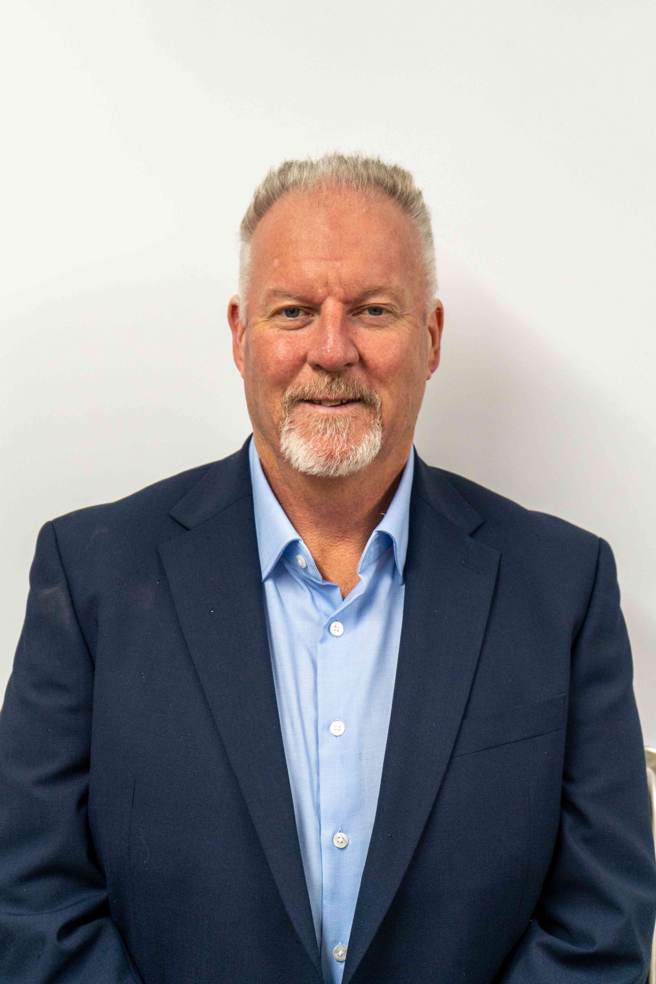 A middle-aged man with short gray hair and a goatee, wearing a navy blazer and light blue dress shirt, standing against a plain white background.