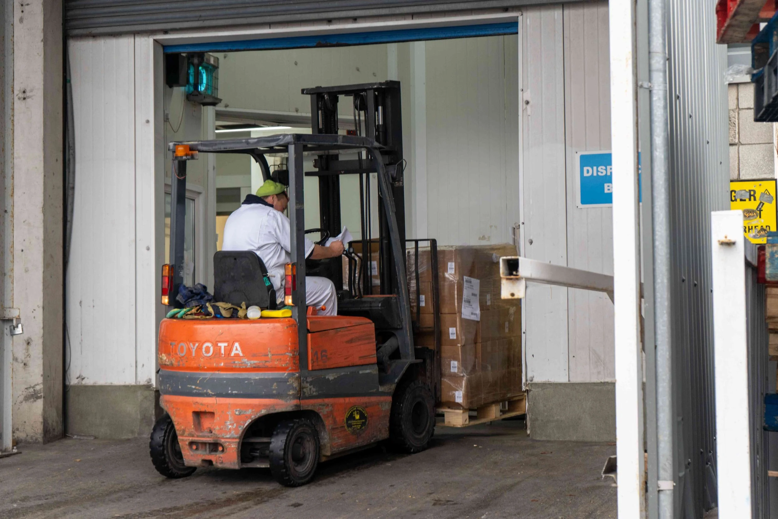 A worker operating an orange Toyota forklift inside a warehouse or storage facility, lifting a pallet of cardboard boxes.