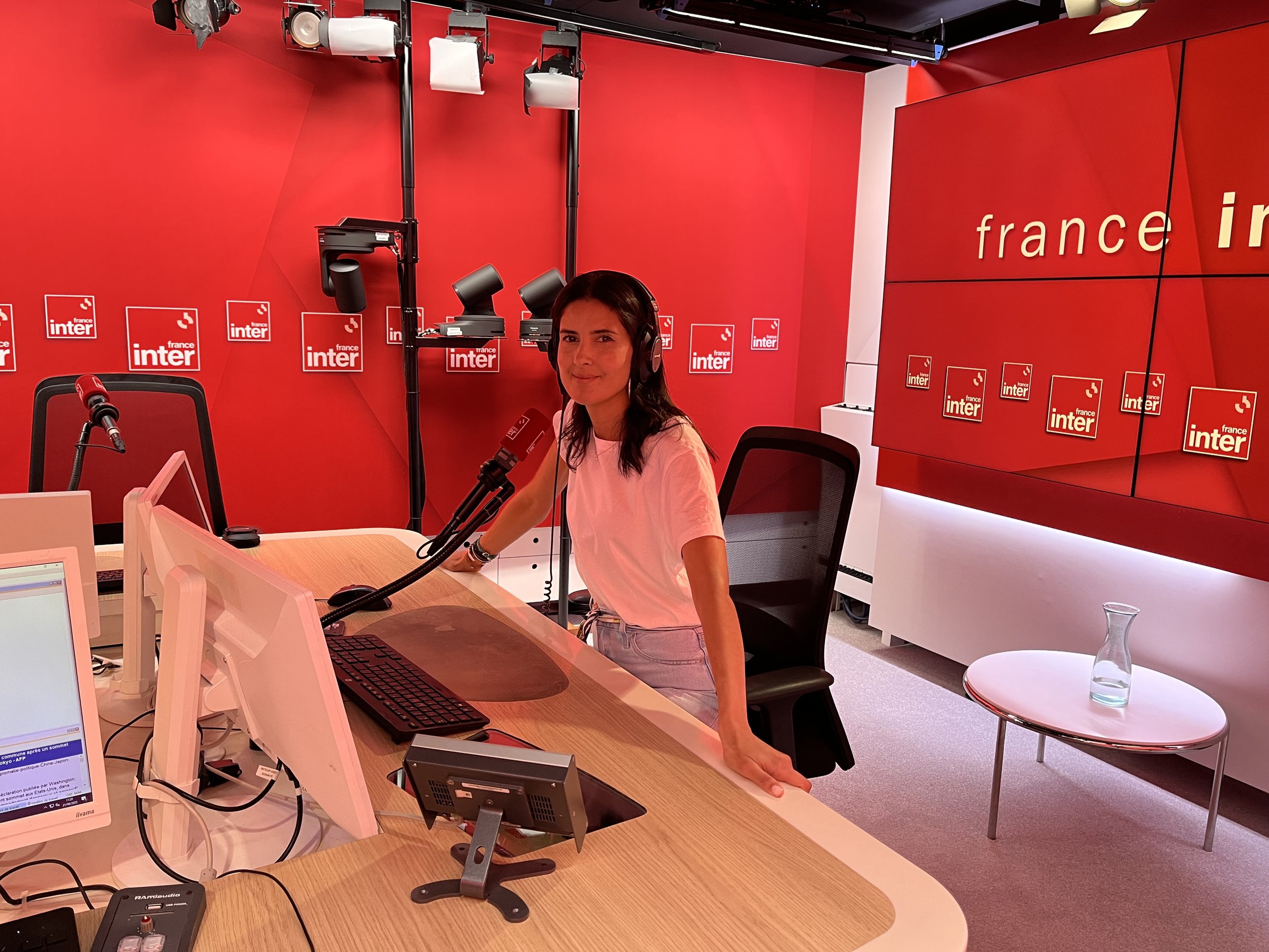 Une femme dans un studio de radio, portant un casque, assise à une table avec un micro, entourée d'équipements de studio, avec des murs rouges et le logo 'France Inter' en arrière-plan.