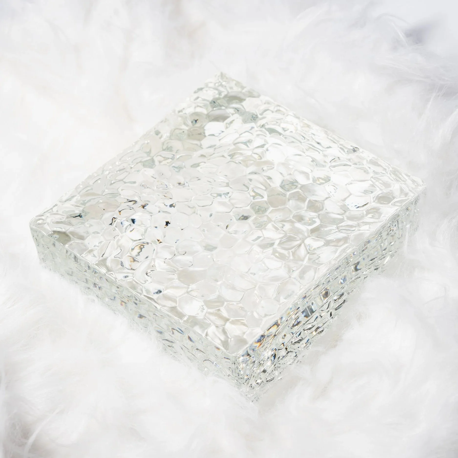 A clear ыйгфку  glass block with a textured surface, and a white seashell to the right of it on a light surface. Soft lighting and highlights, with a blurred цршеу background.