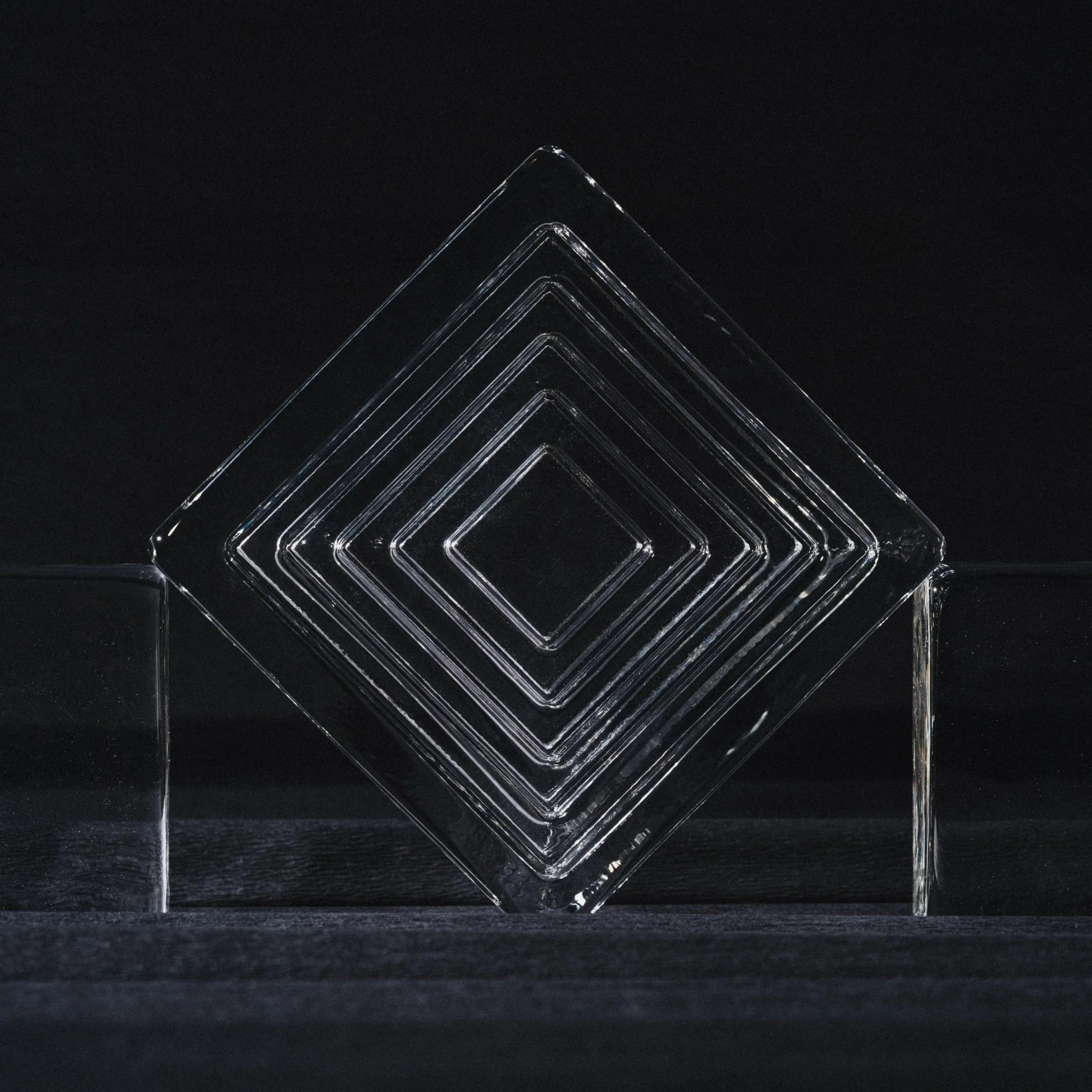 A rectangular solid glass block with a metallic textured surface, placed on a shimmery, black, reflective surface.