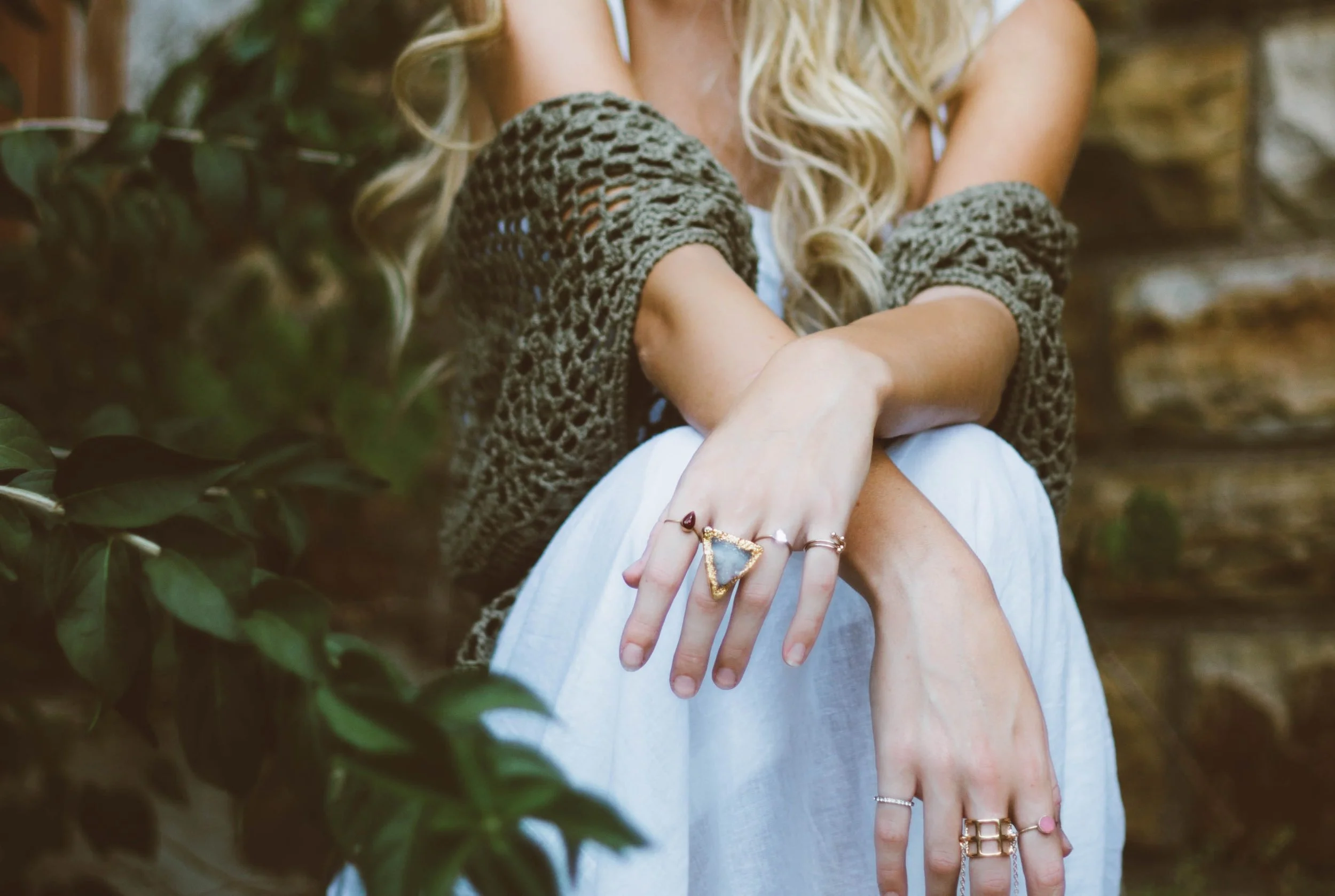 Femme avec cheveux blonds longs portant une veste en tricot gris et des bracelets, montrant ses mains et ses doigts garnis de bijoux, en arrière-plan de verdure.