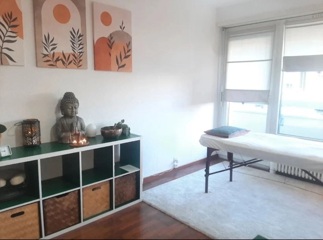 Salle de massage avec un lit de massage, une étagère verte avec des décorations, une statuette de Bouddha, des bougies, une peinture abstraite sur le mur avec des couleurs pastel, un tapis beige, et une grande fenêtre avec volets.