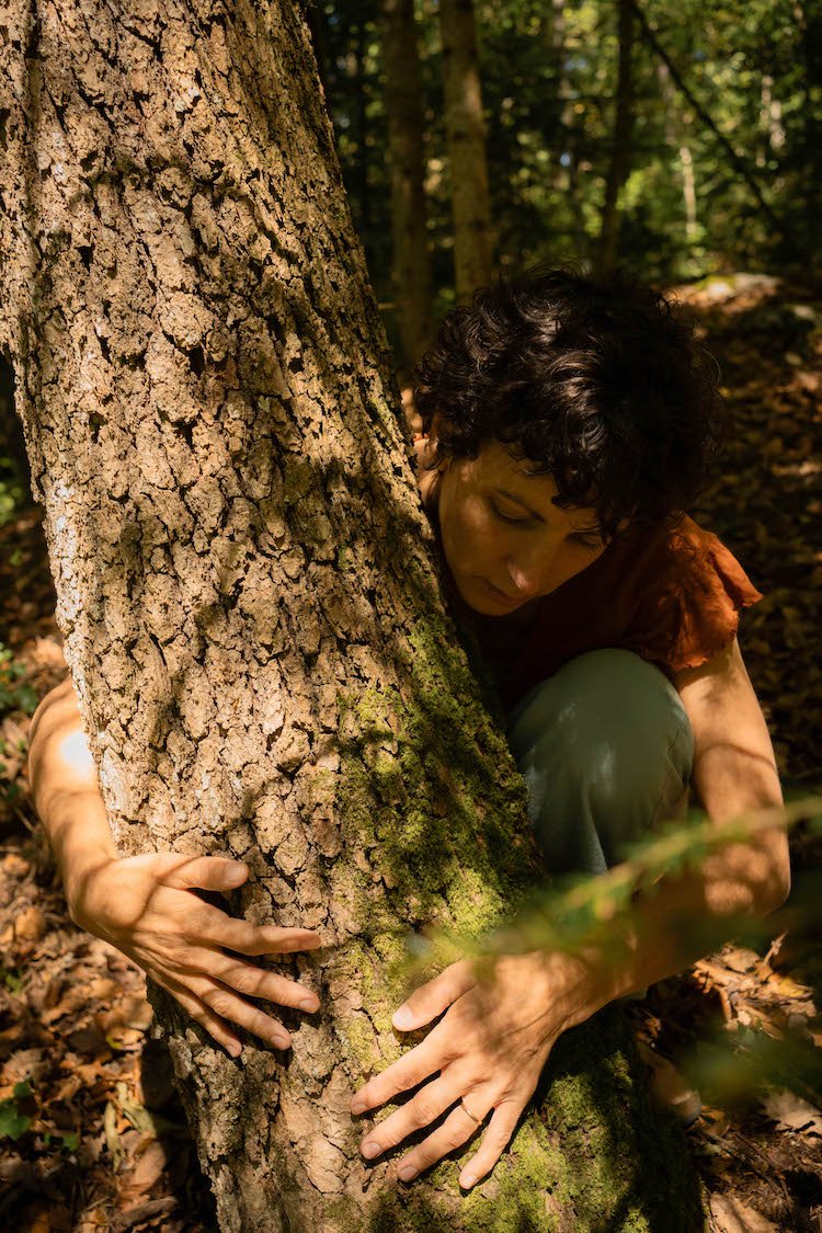 Une femme avec des cheveux bouclés fait semblant d'embrasser ou de faire semblant d'étreindre un arbre dans une forêt.