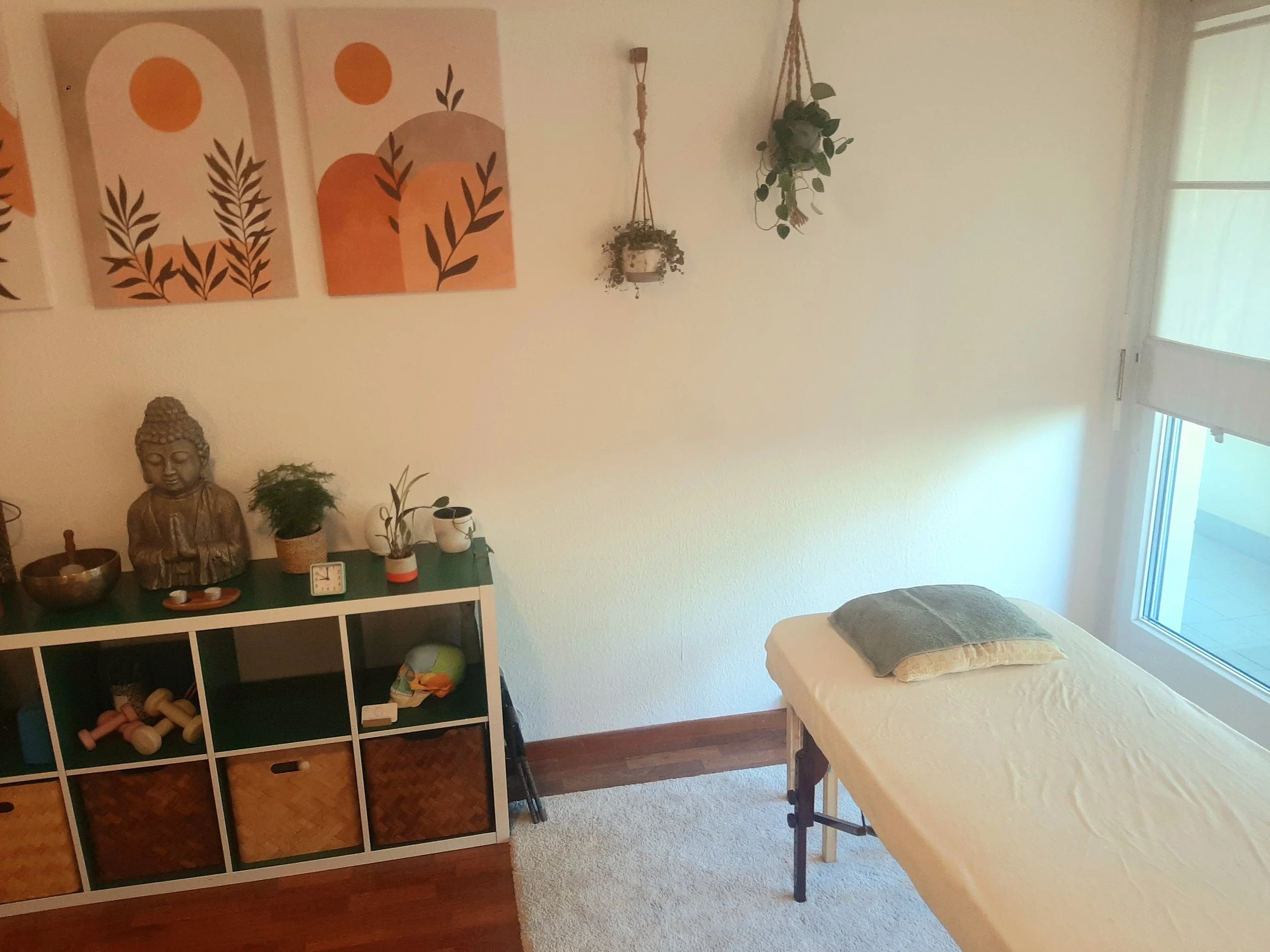 Une pièce de chambre ou salon avec quelques décorations sur le mur, un meuble avec des objets de décoration, une statue de Bouddha, et un lit avec un oreiller. La pièce est bien éclairée par une grande fenêtre.