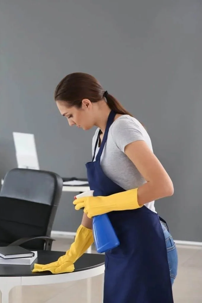 Une femme en tenue de nettoyage, portant des gants jaunes, nettoie une table de bureau avec un spray, dans un bureau moderne.