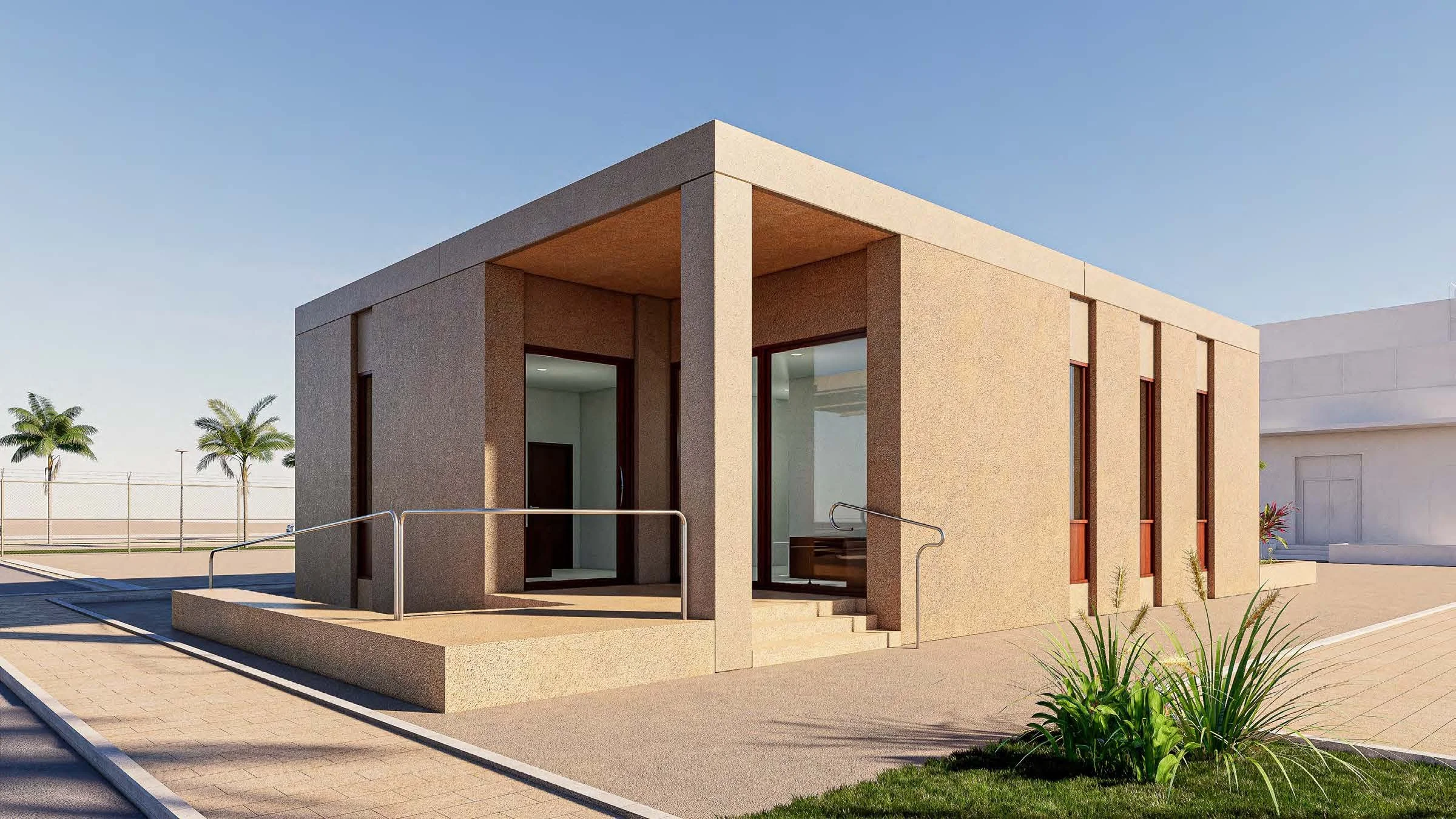 Vista frontal de un edificio moderno de una planta, con paredes de piedra y ventanas largas y estrechas, rodeado de un área pavimentada y áreas verdes con plantas y palmeras en el fondo.