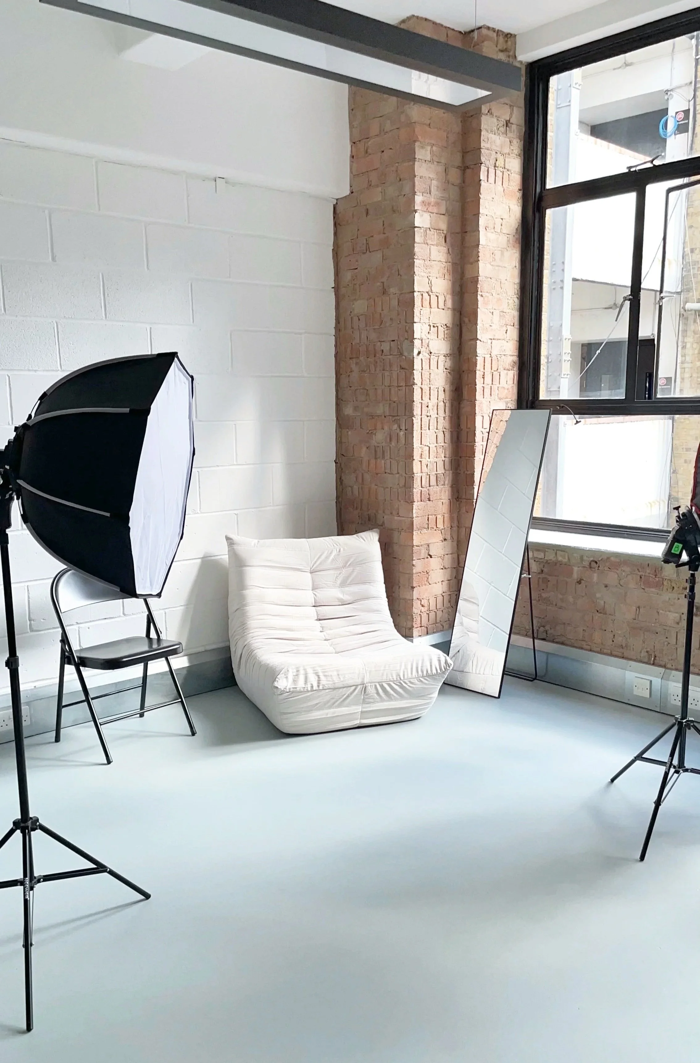 An indoor photography studio with a white brick wall, a large window, studio lighting equipment, a white cushioned chair, and a full-length mirror.