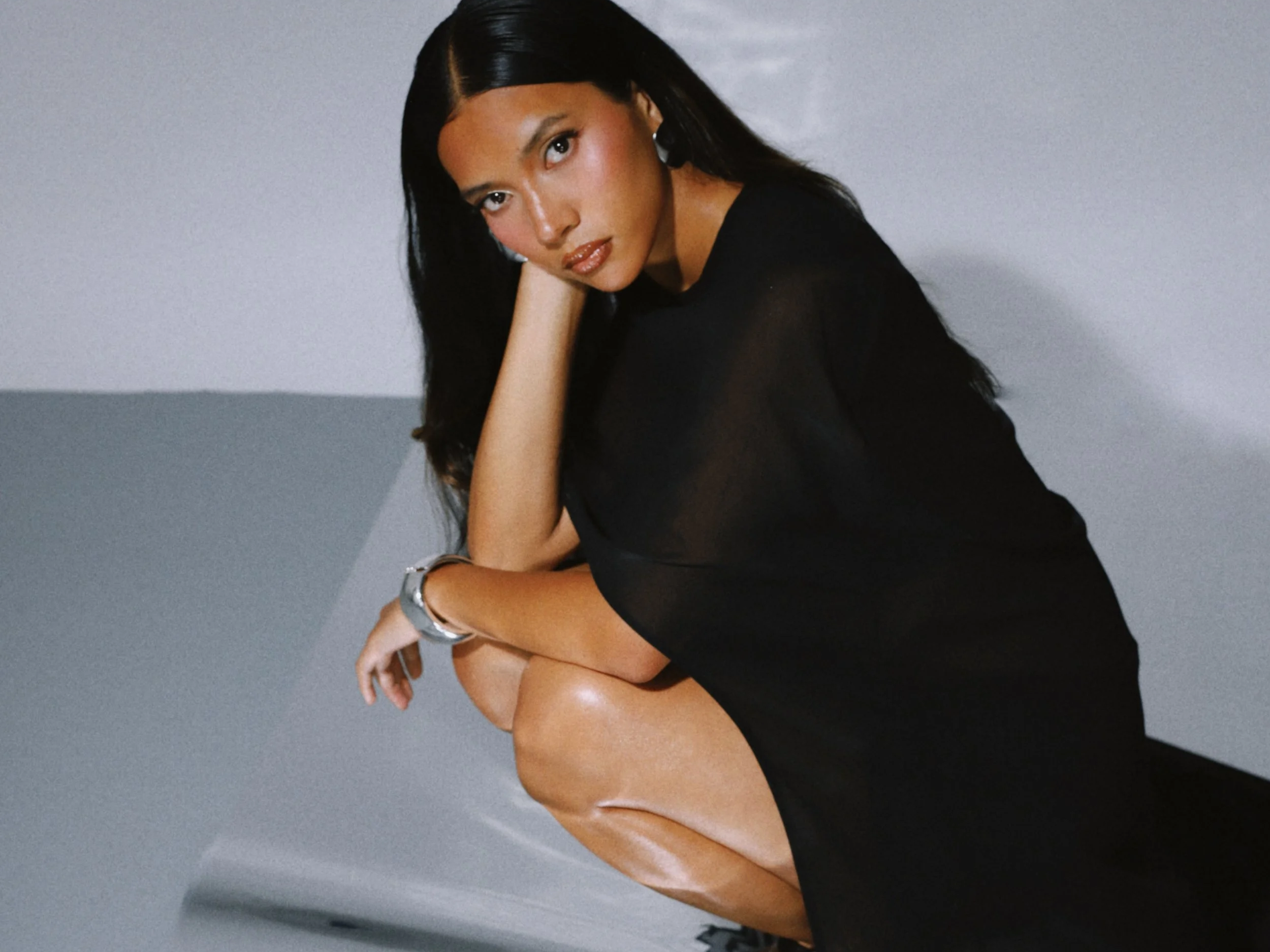 A woman with long dark hair and tan skin is seated with her head resting on her knee. She is wearing a black sheer top and has a silver watch on her wrist. The background is plain and neutral, possibly a gray or white wall.