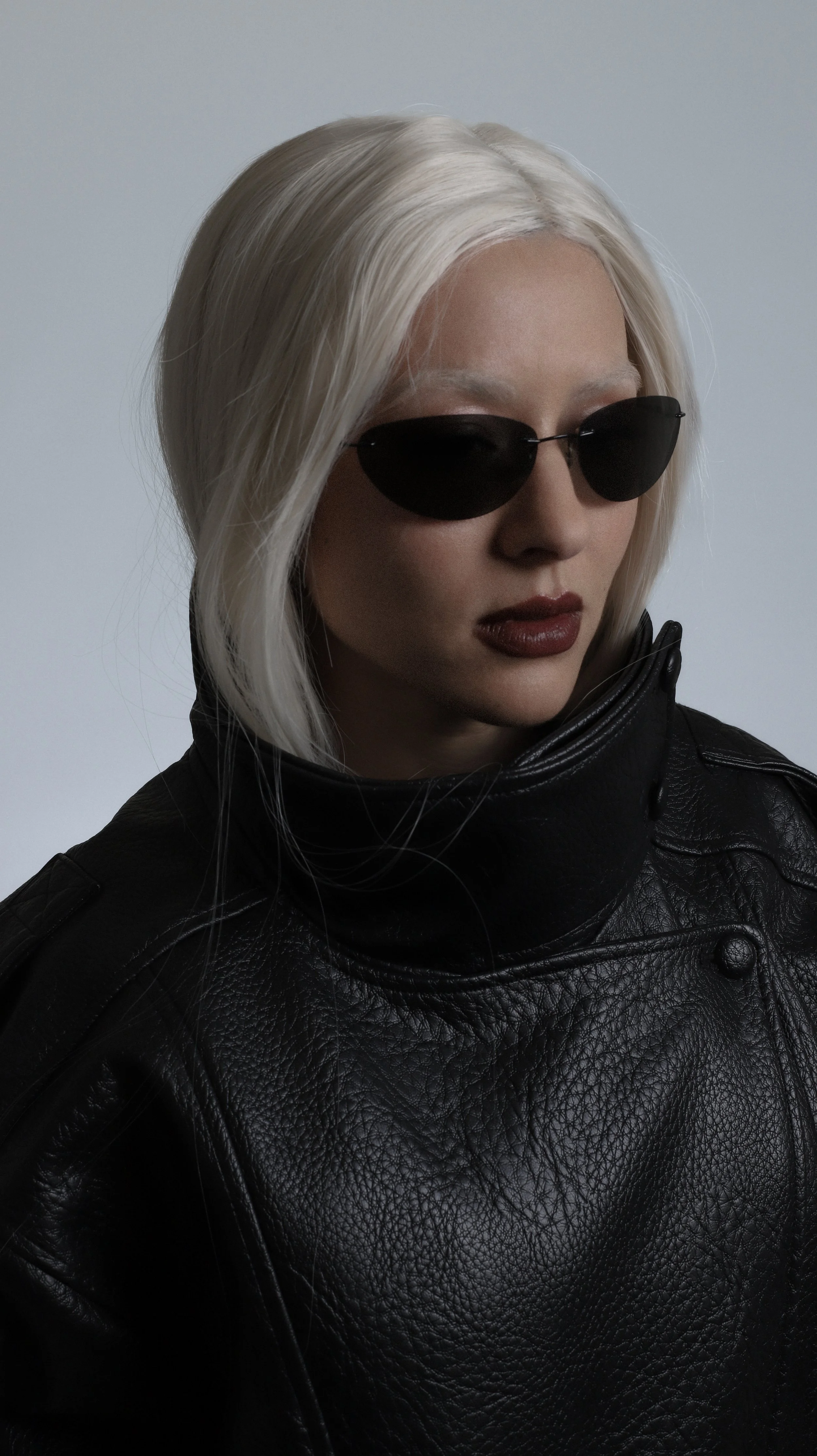 A woman with platinum blonde hair wearing black sunglasses and a black leather jacket against a plain background.
