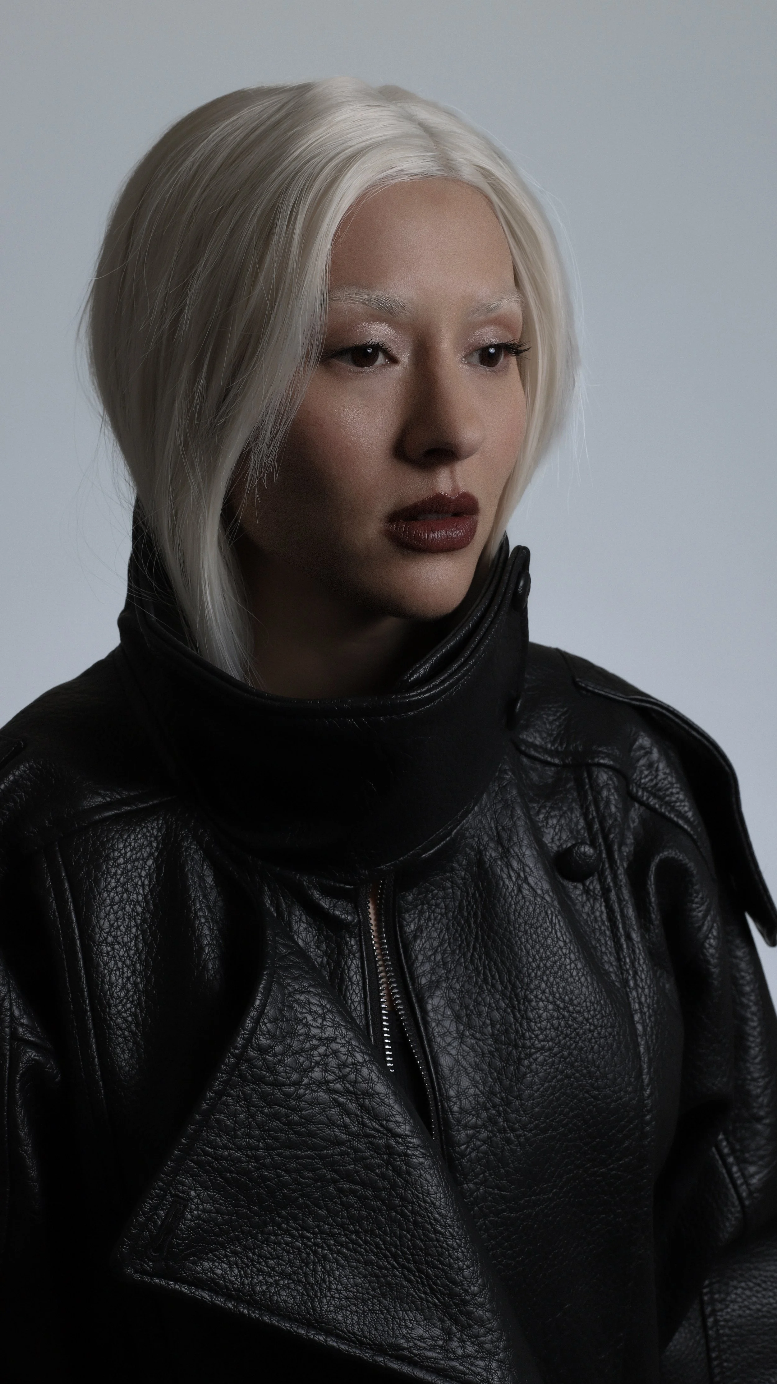 A young woman with platinum blonde hair and fair skin, wearing a black leather jacket, looking contemplative or serious.