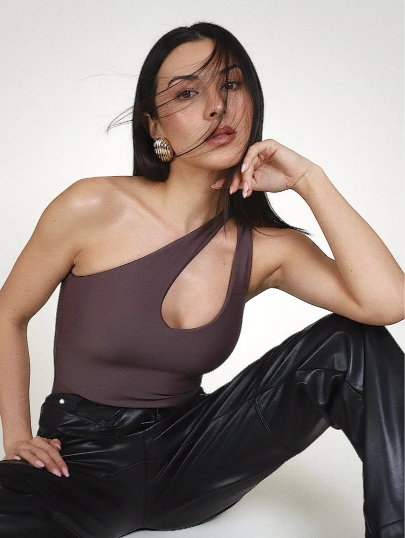 A woman with long black hair and large earrings posing against a plain background. She is wearing a one-shoulder, cut-out top and black leather pants.