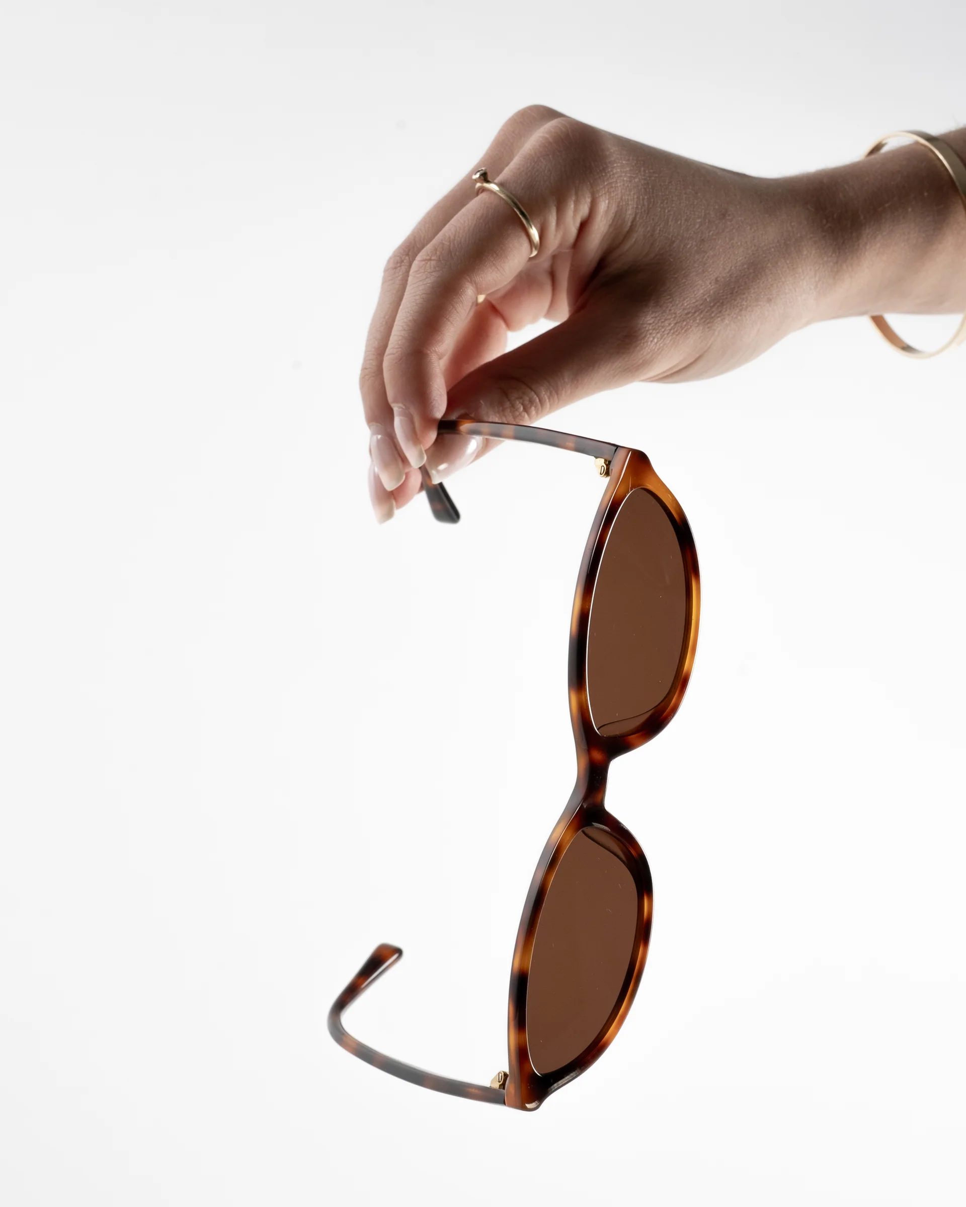 A person's hand holding a pair of tortoise shell sunglasses against a white background.