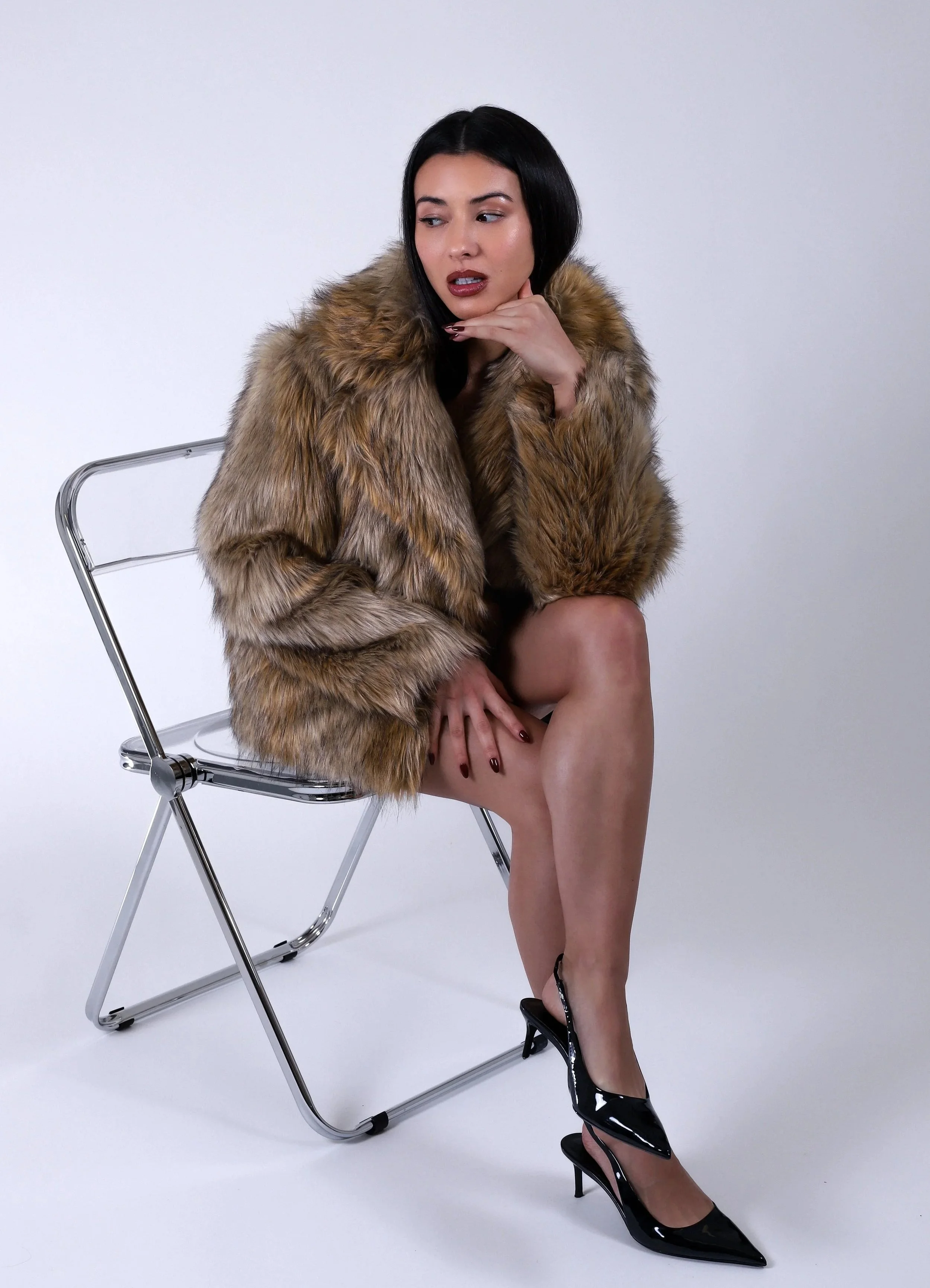 A woman with dark hair wearing a fur coat, sitting on a metal folding chair, with one leg crossed over the other, wearing black high heels, posing against a plain white background.