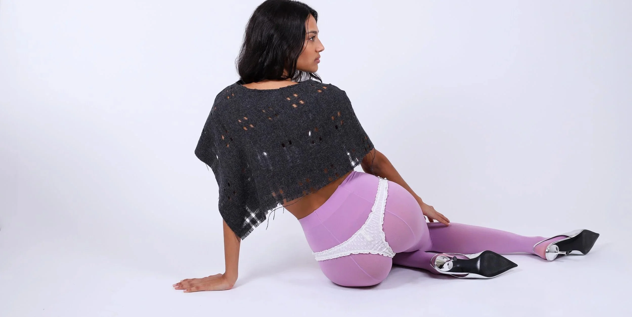 A woman in light purple tights and distinctive high-heeled shoes is sitting on the floor with her back and side to the camera, in front of a plain white background.