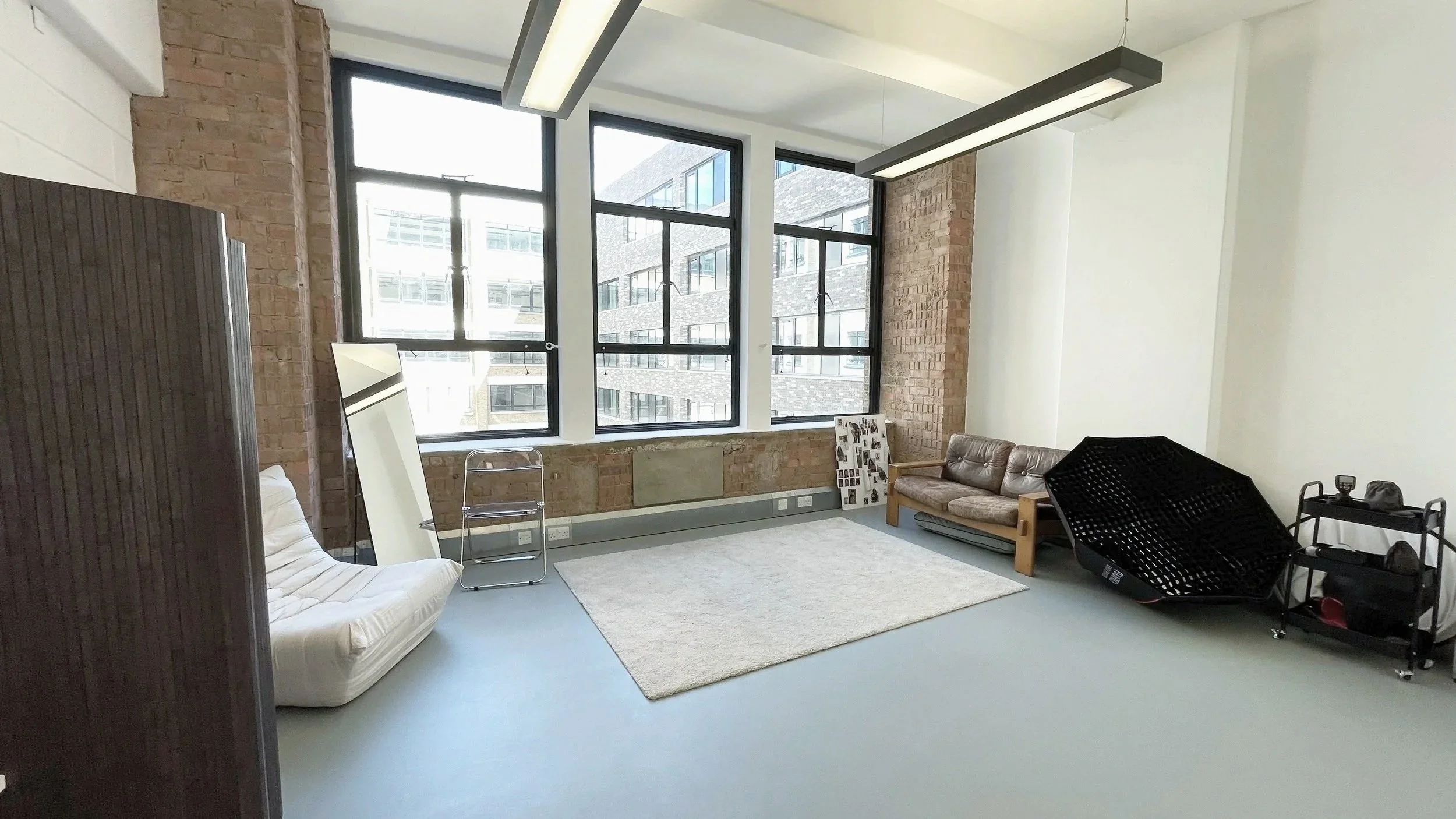 Industrial-style office space with large windows, brick walls, a beige sofa, a white armchair, laser printer, photo collage, and black chair.