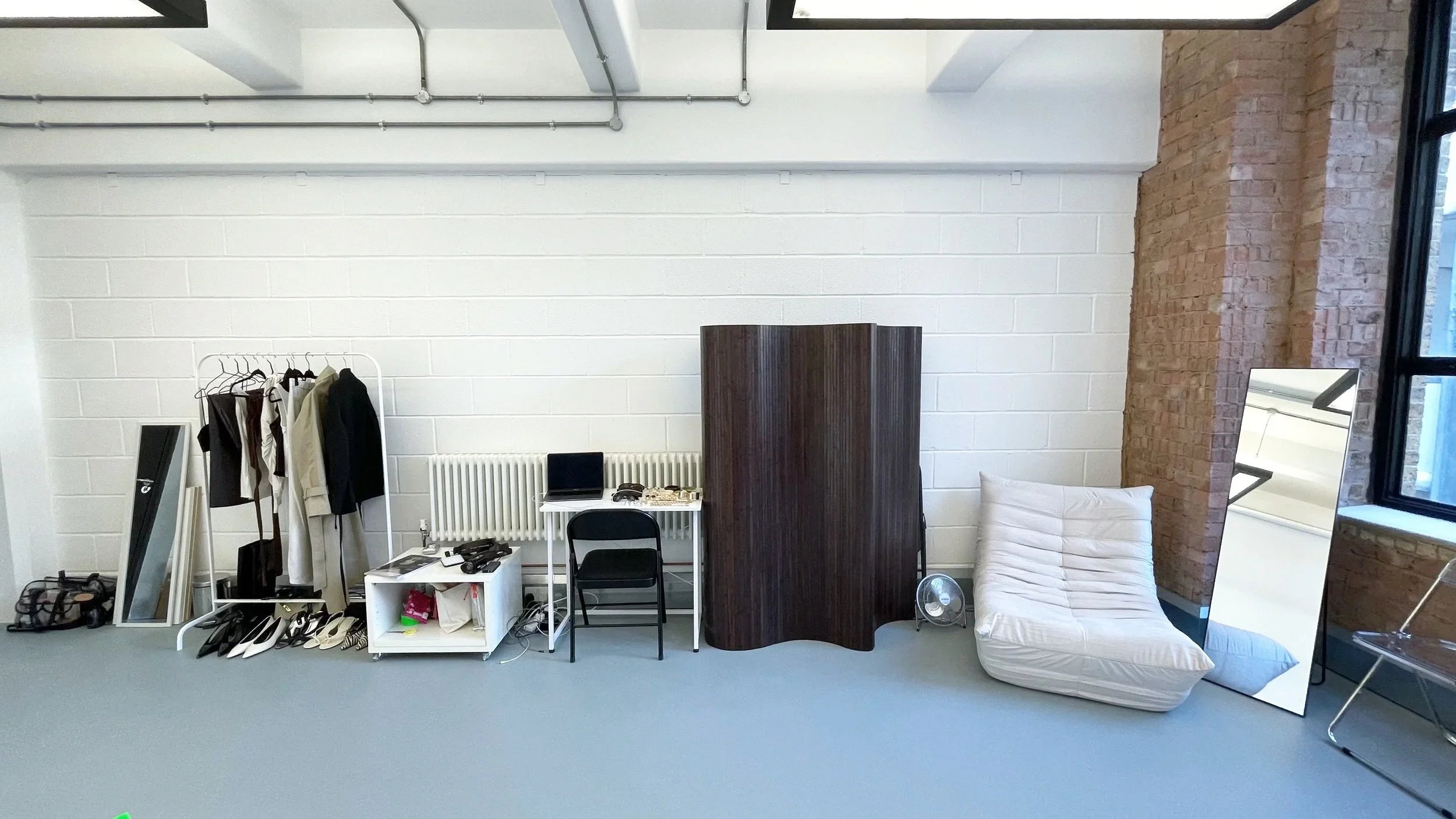 An interior space with a white cinder block wall, exposed brick column, window, mirror, a white chair with a pillow, a small table, a clothing rack with clothes, and various objects on the floor and furniture.