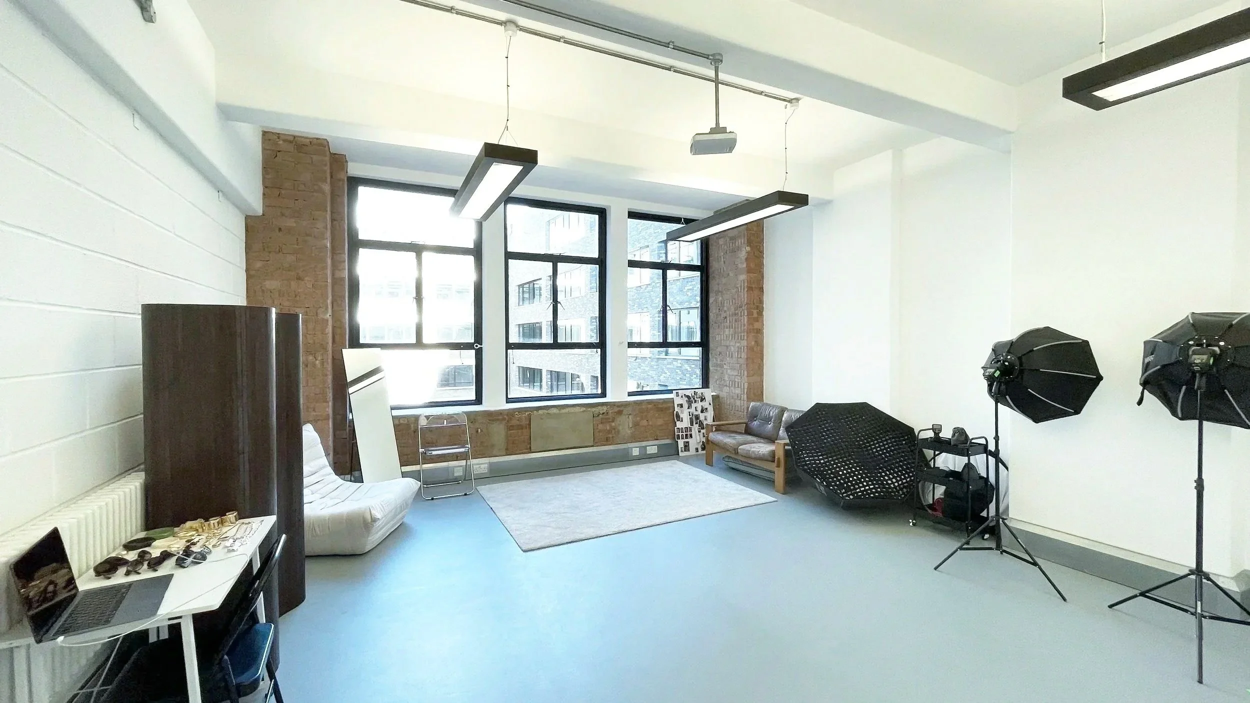 A photography studio with large windows letting in natural light, white and brick walls, and various lighting equipment including softboxes. There's a small table with accessories, a laptop on a white table, a laptop, a mirror, a white sofa, a bench,