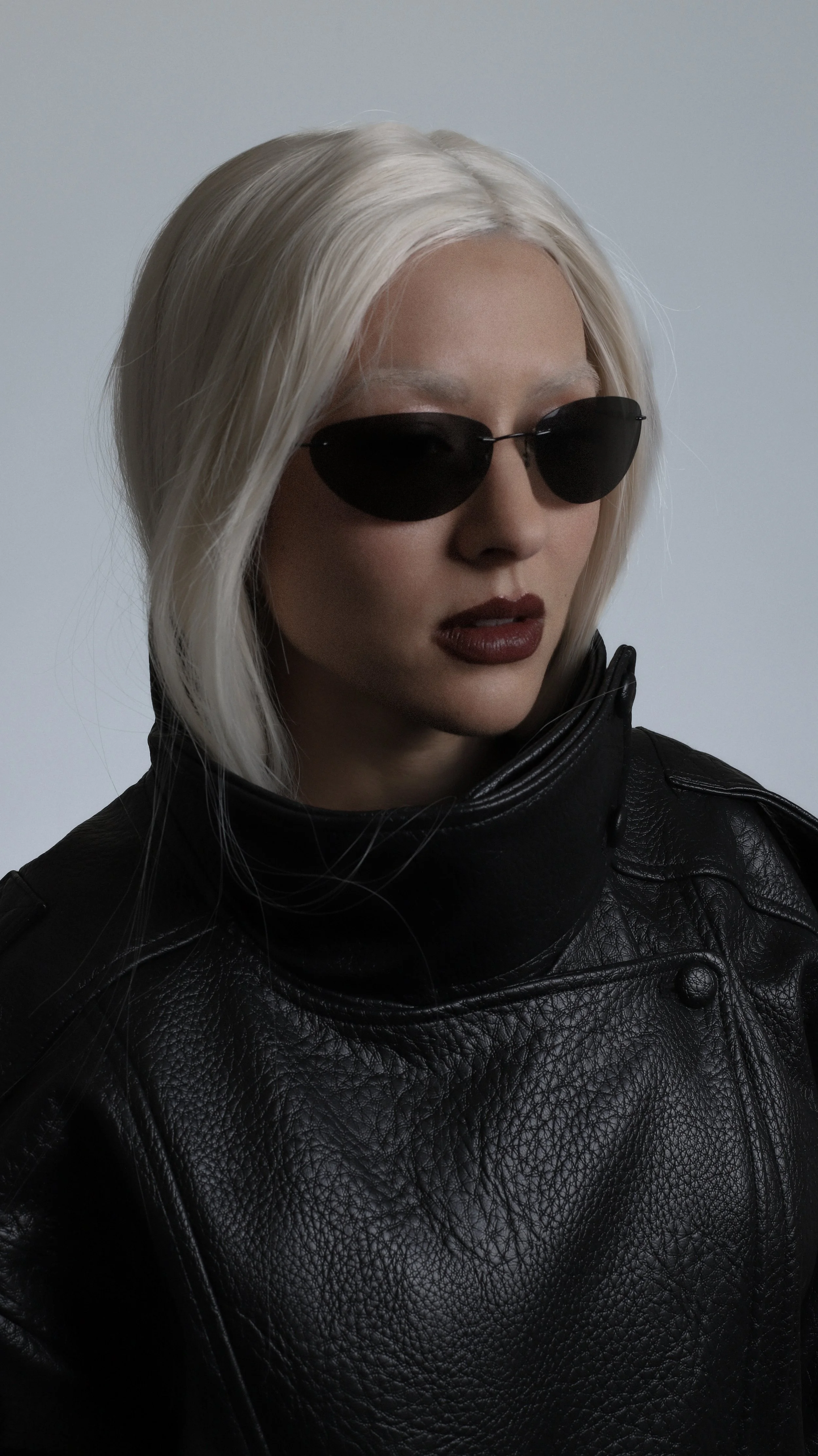 A woman with platinum blonde hair wearing black sunglasses, dark lipstick, and a black leather jacket.