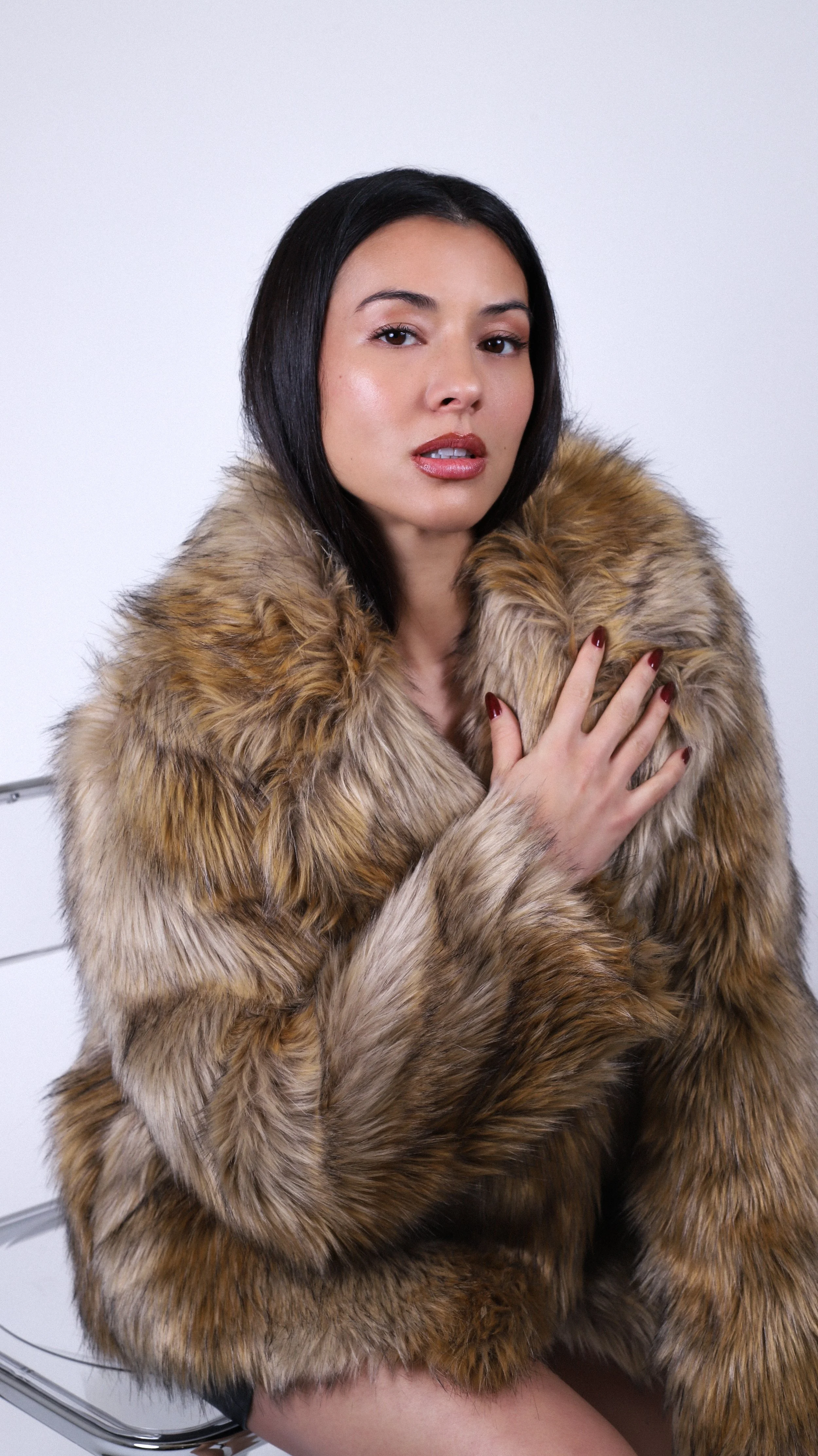A woman with dark hair wearing a fur coat, sitting on a chrome chair, holding her chest with one hand, against a plain white background.