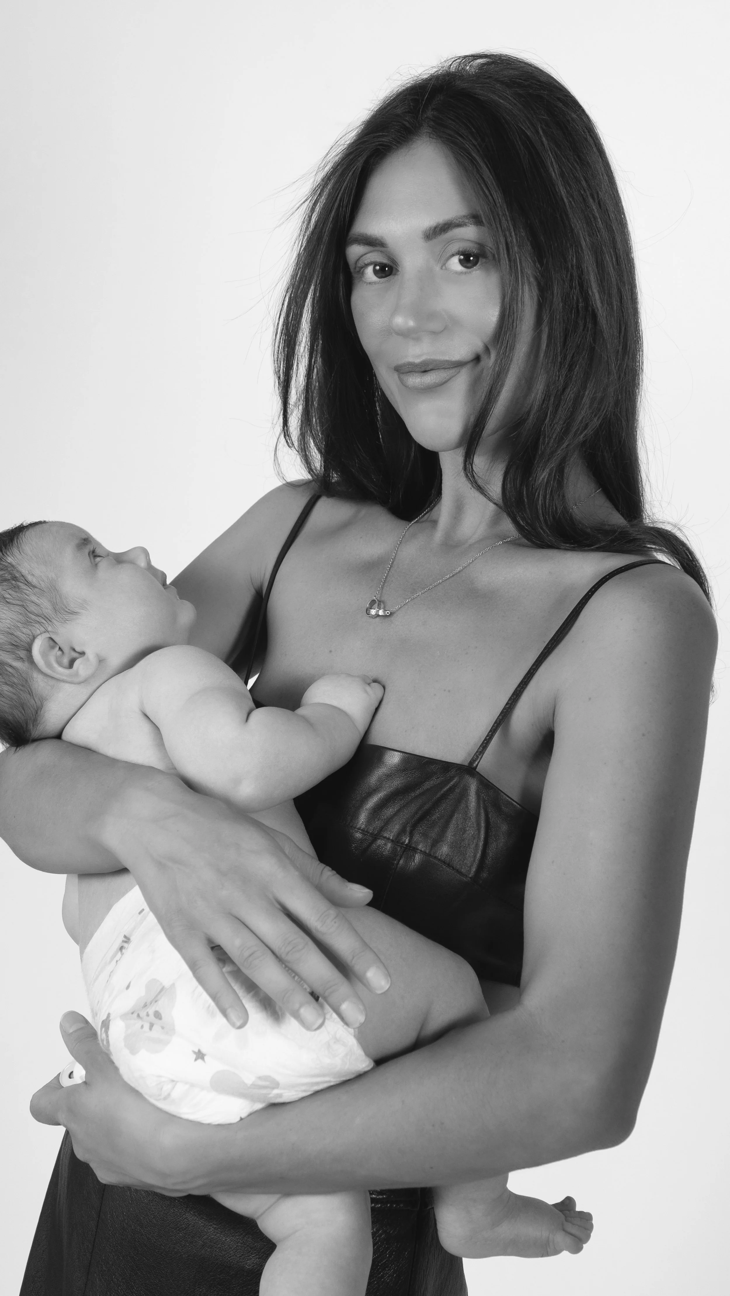 A woman with long dark hair holding a baby in her arms, both looking at each other. The woman is smiling slightly and wearing a black leather top. The baby is looking up at the woman and is dressed in a diaper with colorful star patterns. The backgro