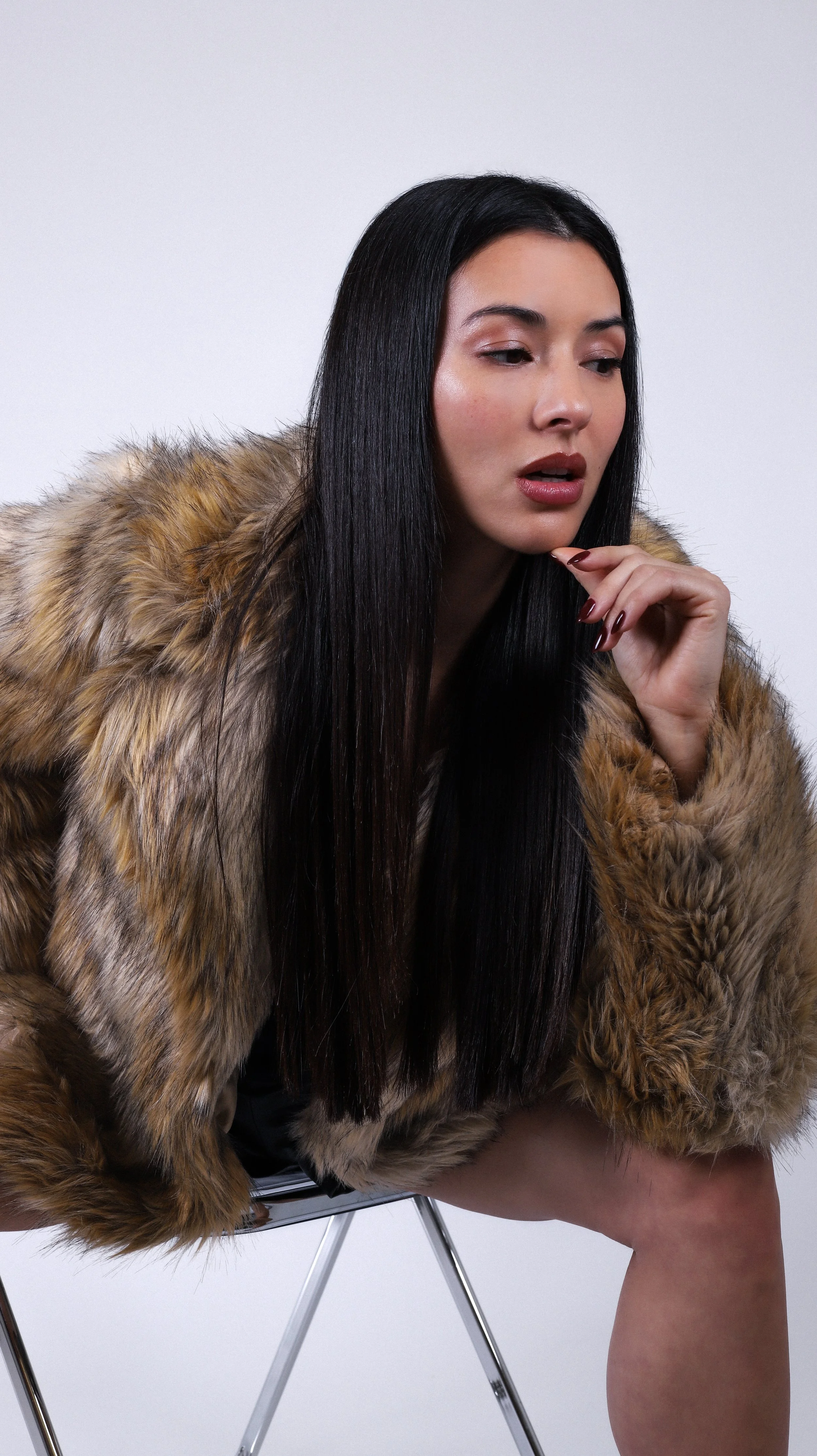 A woman with long black hair wearing a fur coat sitting on a modern stool against a plain white background.