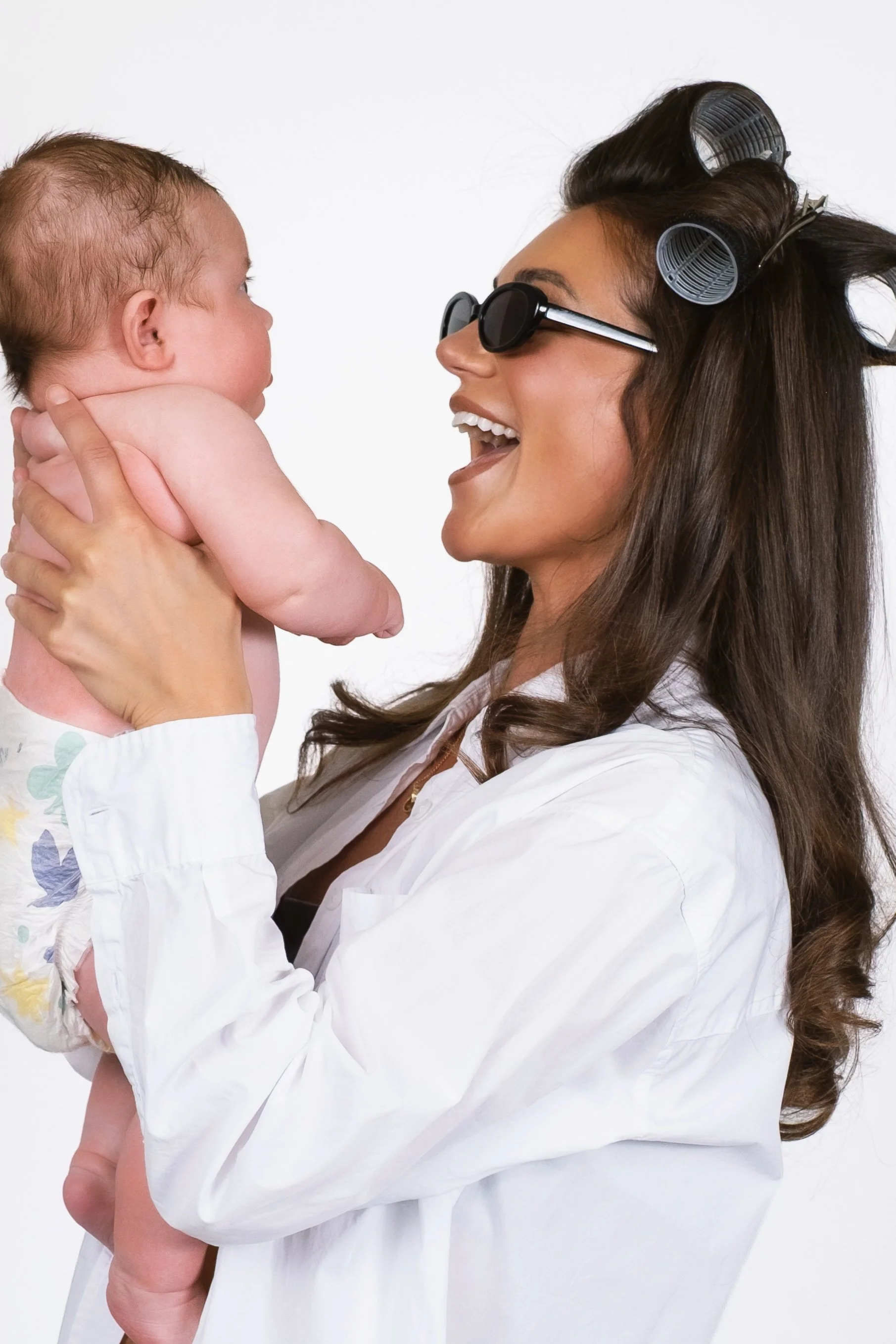 Woman wearing black sunglasses and hair rollers holding a baby, who is shirtless and looking at her